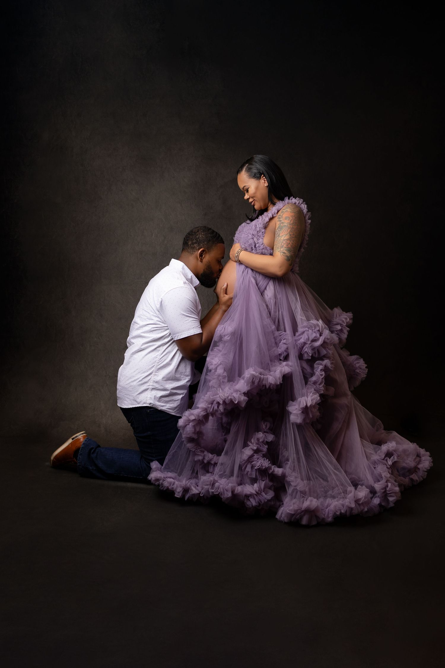 Man kneeling and kissing pregnant woman's belly during studio maternity photoshoot in purple gown.