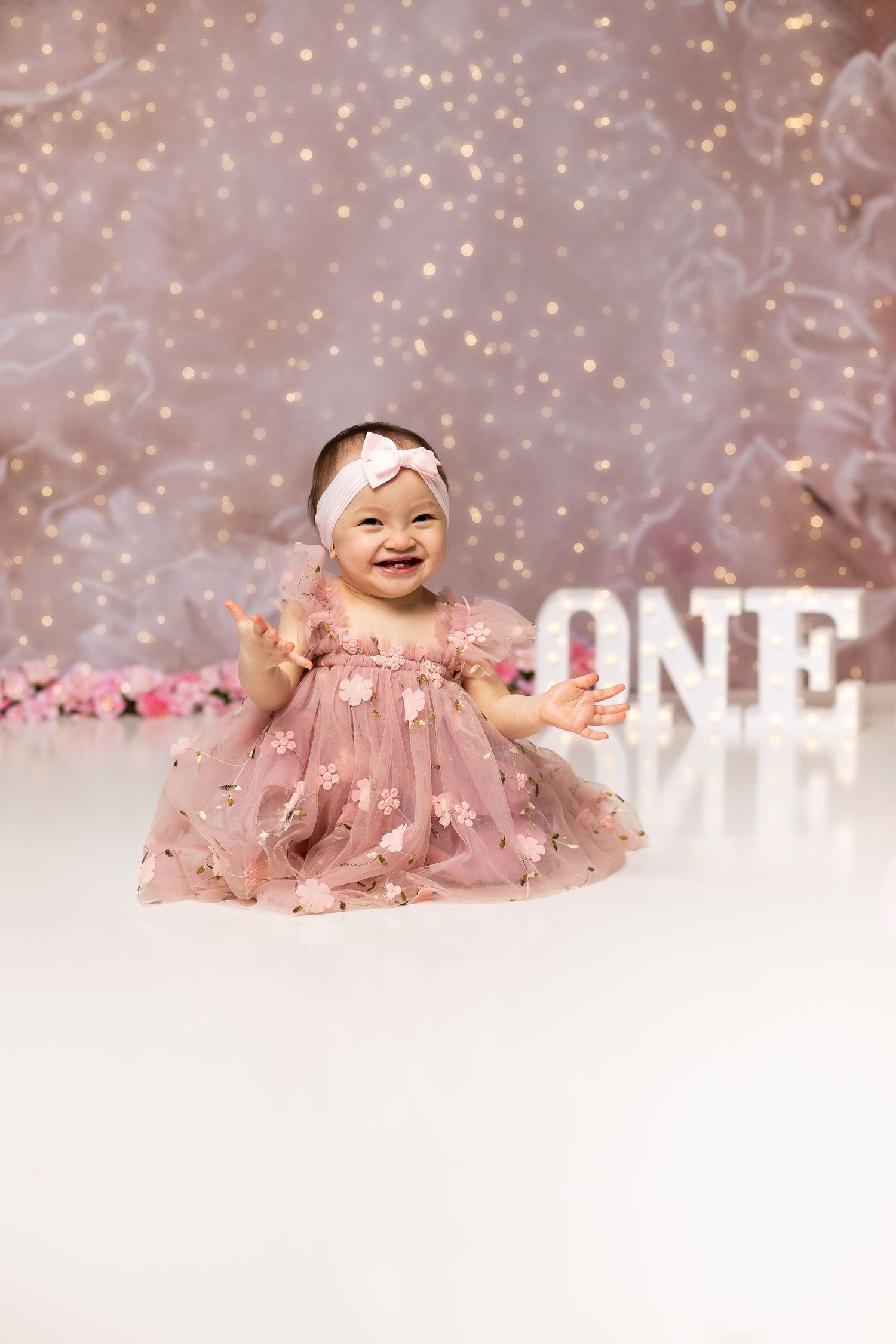 Happy baby girl in floral pink dress laughing beside ONE sign with bokeh light backdrop.