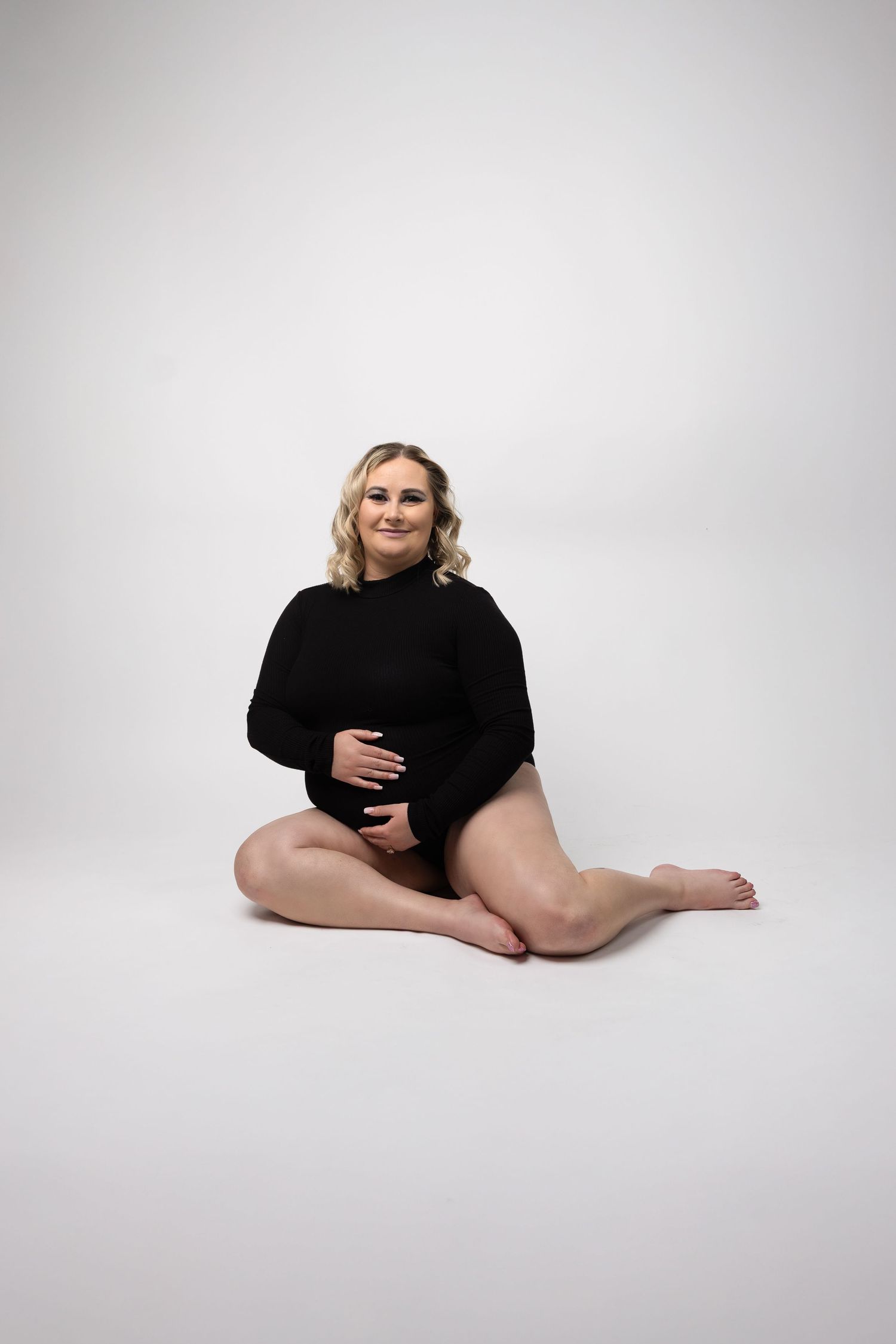 Blonde woman in black bodysuit sits cross-legged on white floor, smiling warmly at camera.