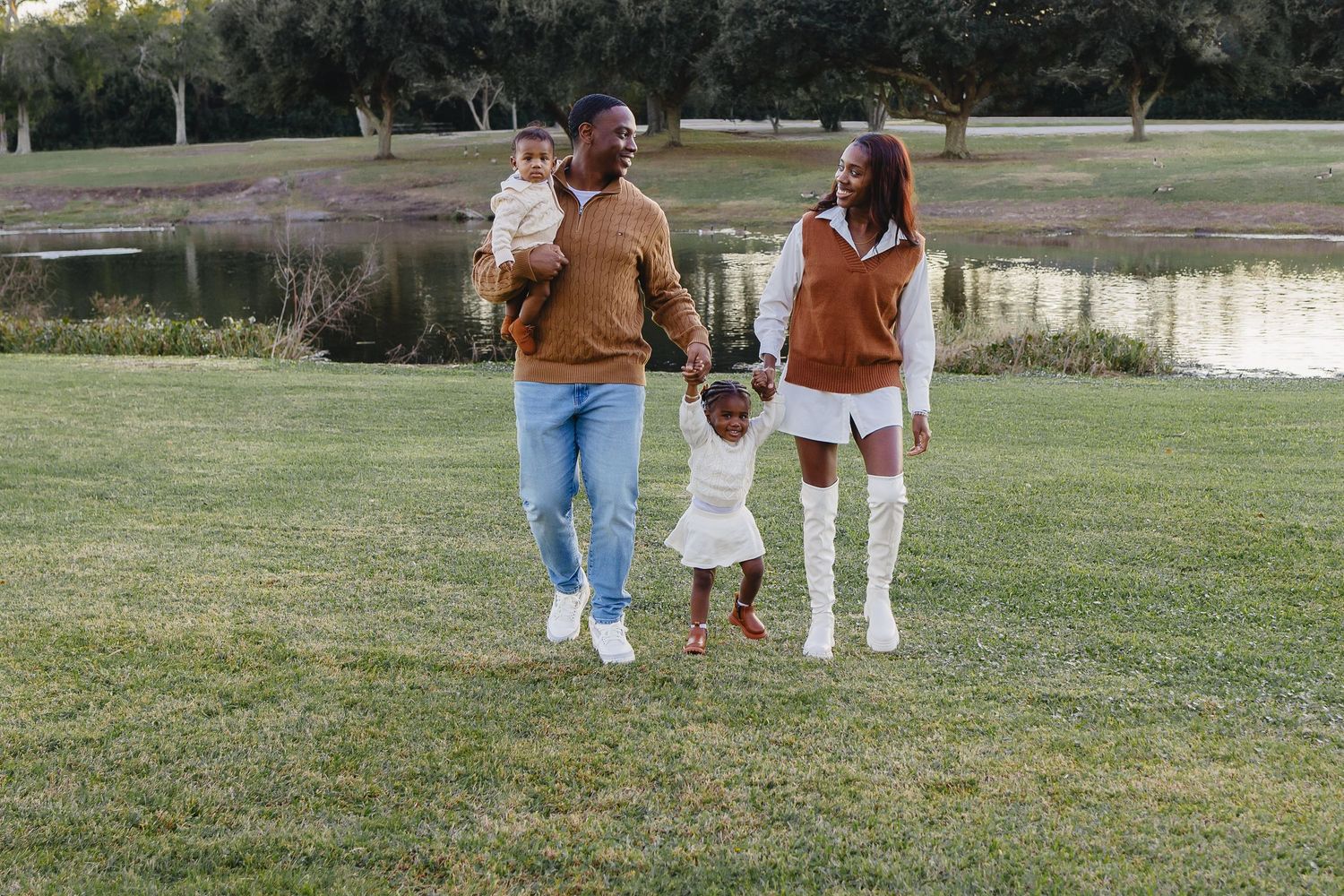 A family of four walking together on a grassy lawn near a peaceful pond at dusk.