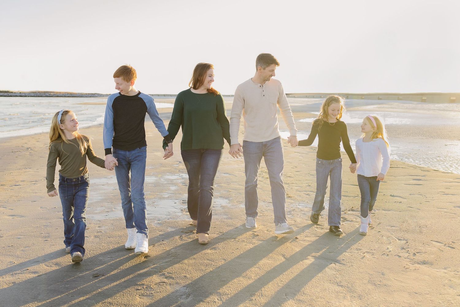 A family of six holds hands and walks along a sunny beach shoreline together.