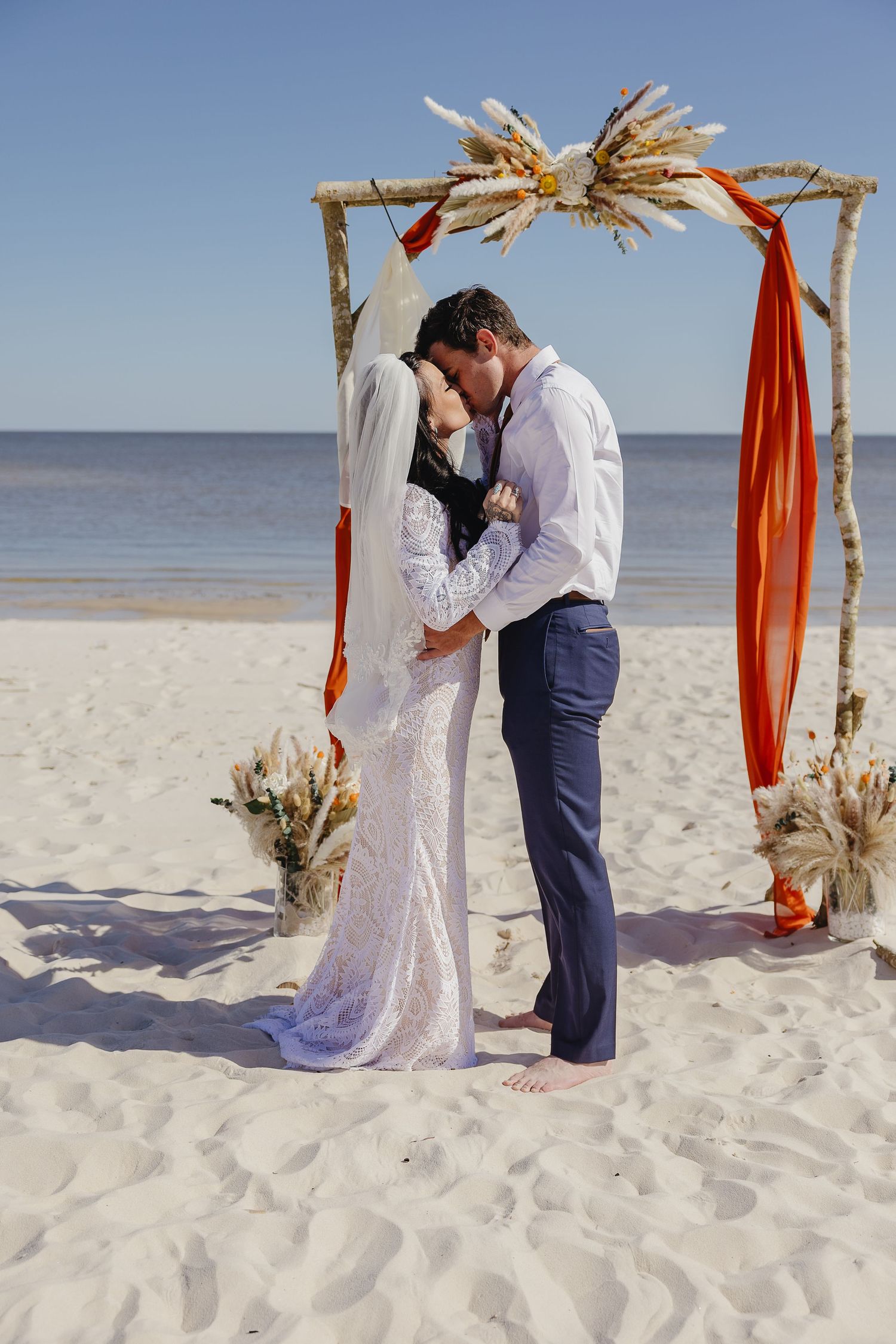 Newlyweds sharing first kiss under boho wooden arch decorated with pampas grass and orange fabric on sandy beach.