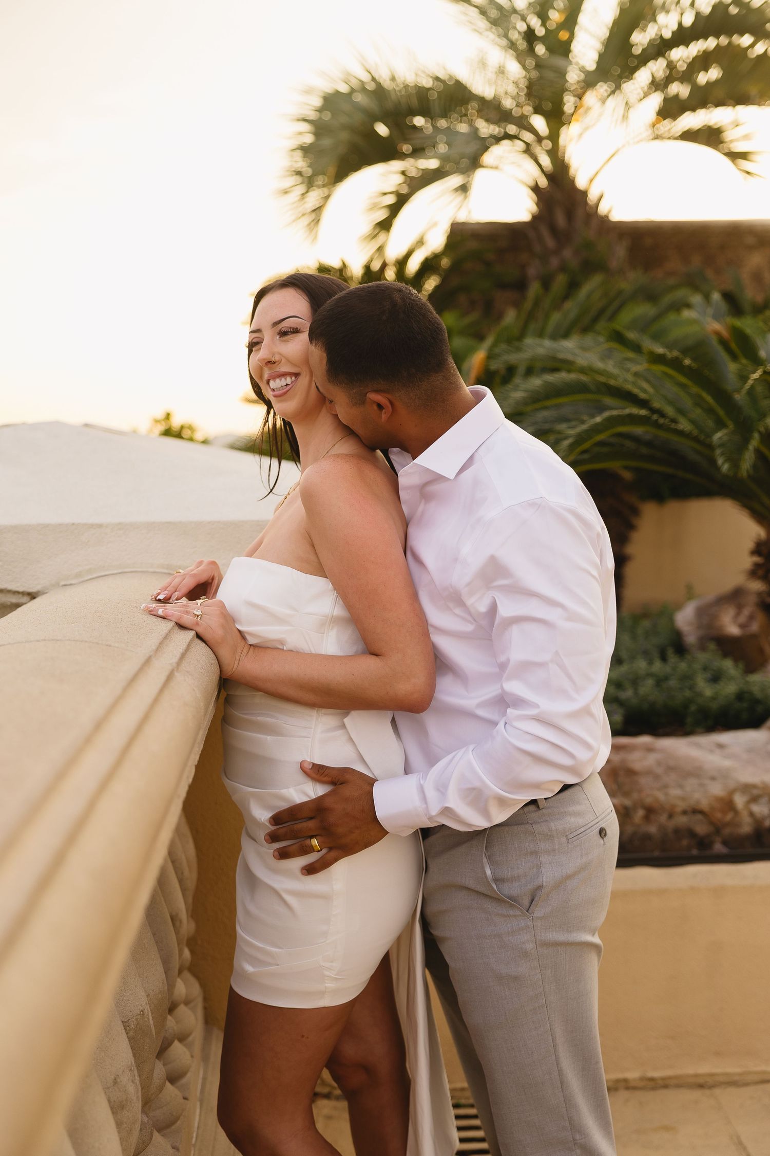 Man in white shirt kissing smiling woman in white dress on outdoor terrace with palm trees at sunset.