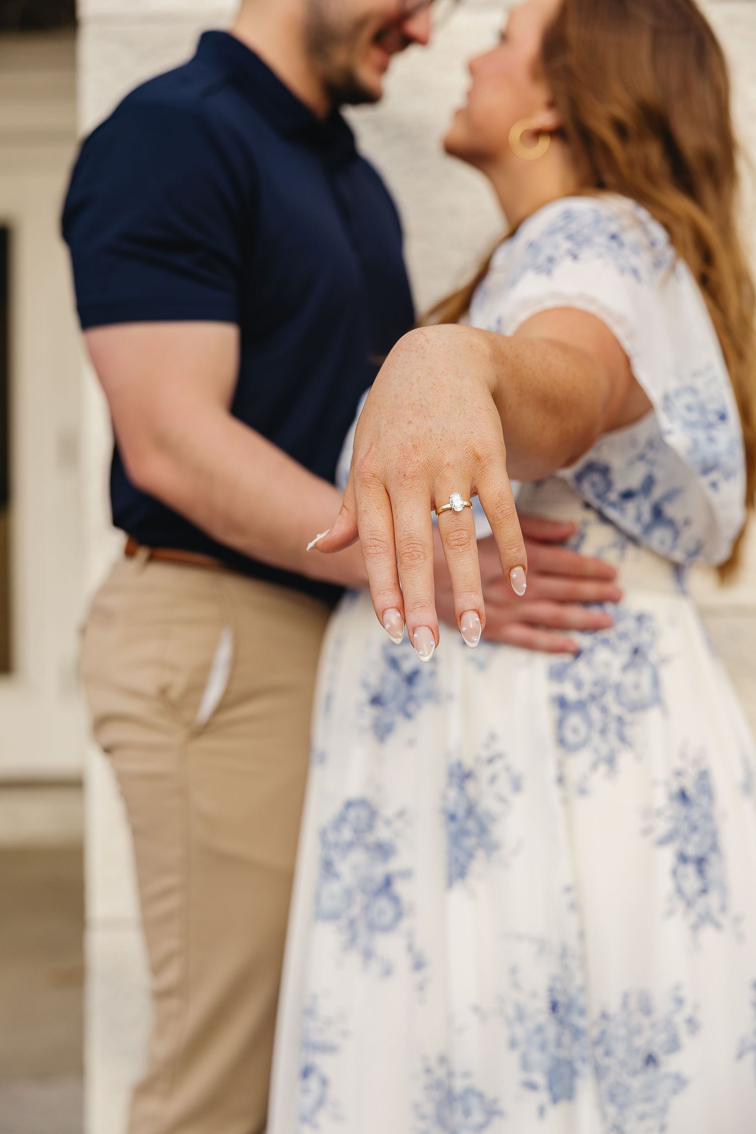 Close-up of womans hand displaying engagement ring while couple embraces, woman in blue floral dress smiling.