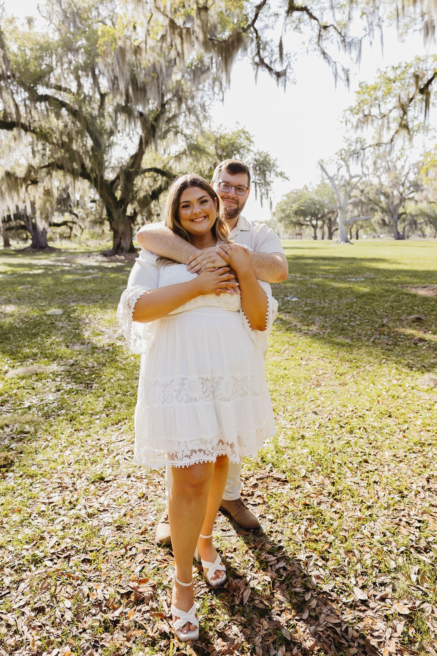 Man embracing woman in white lace dress from behind under Spanish moss oak trees.