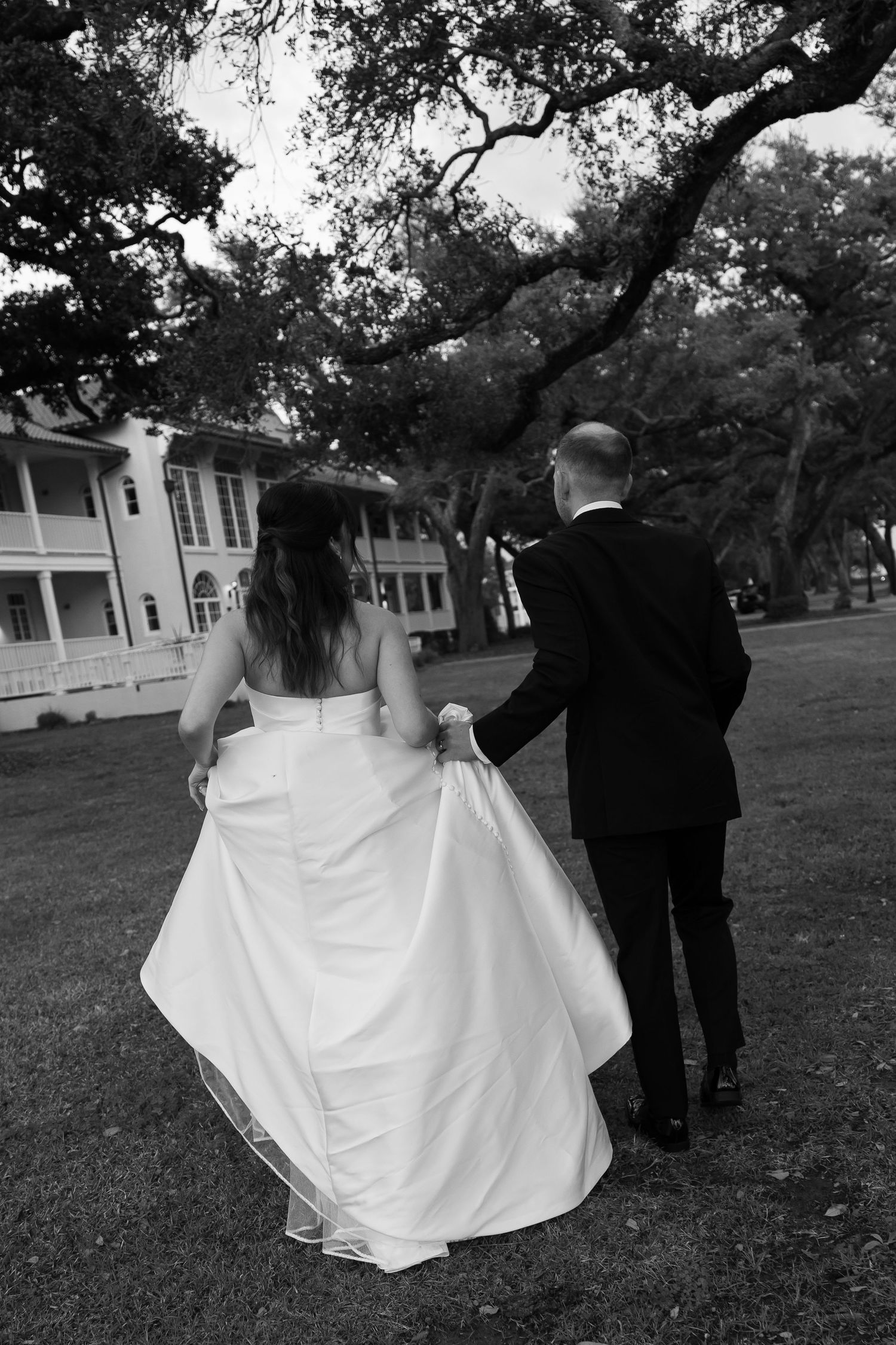 A bride and groom walk hand in hand across a lush lawn in a black and white photo.