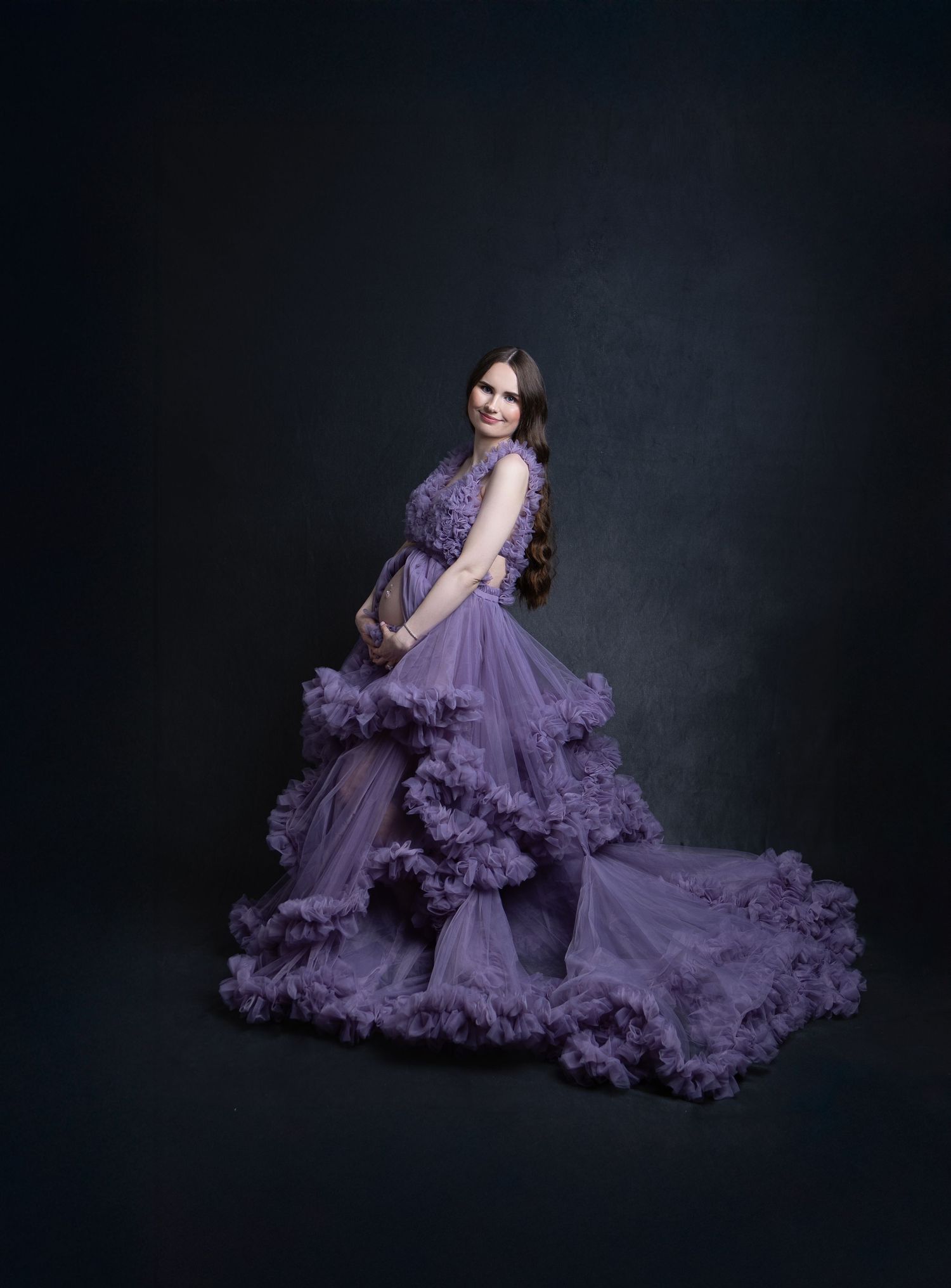 Pregnant woman poses elegantly in a flowing purple ruffled tulle gown against dark backdrop.