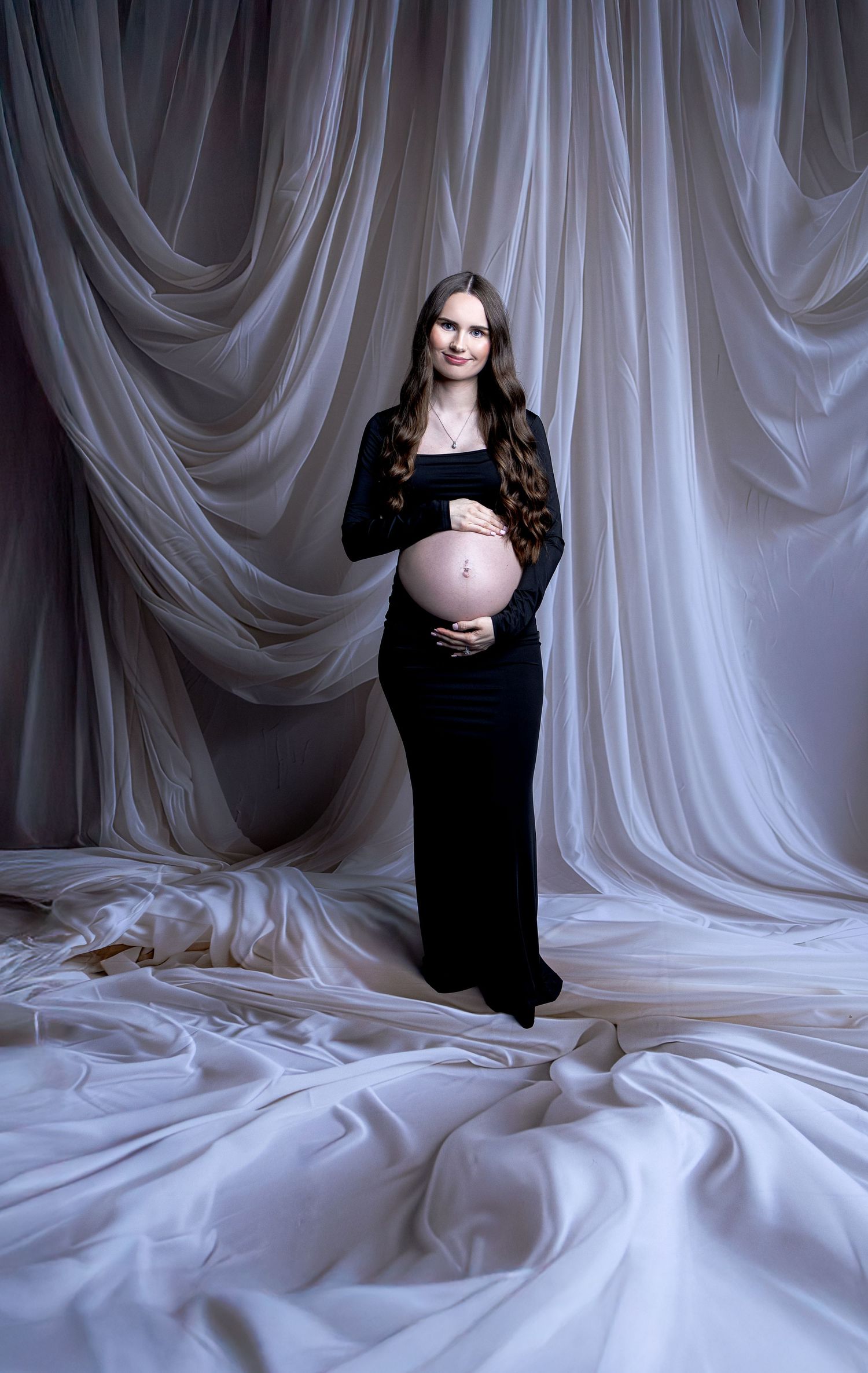 Smiling pregnant woman in black dress posing with draped white fabric studio backdrop.