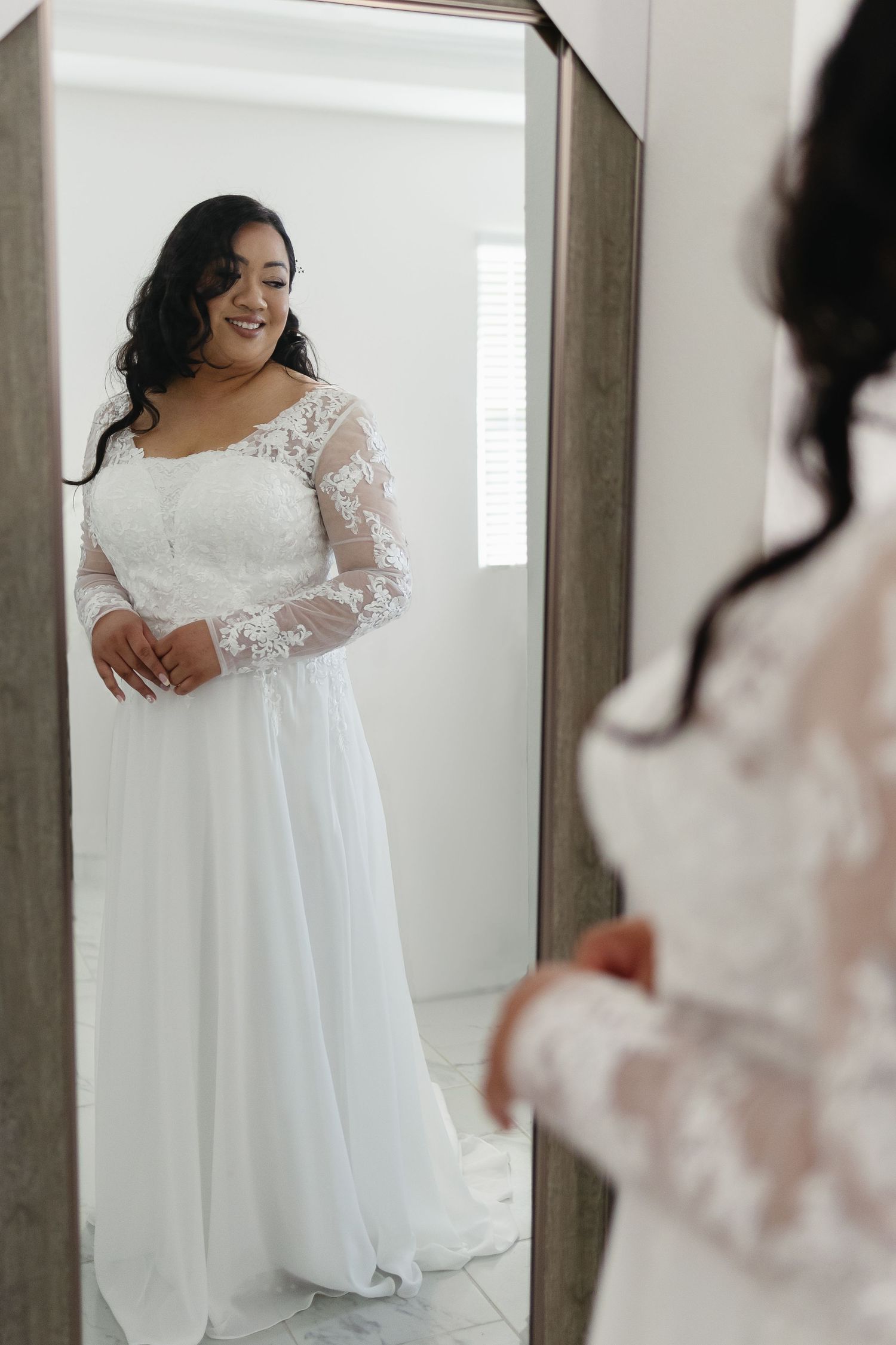Bride in lace long-sleeve wedding gown smiling at her reflection in mirror.