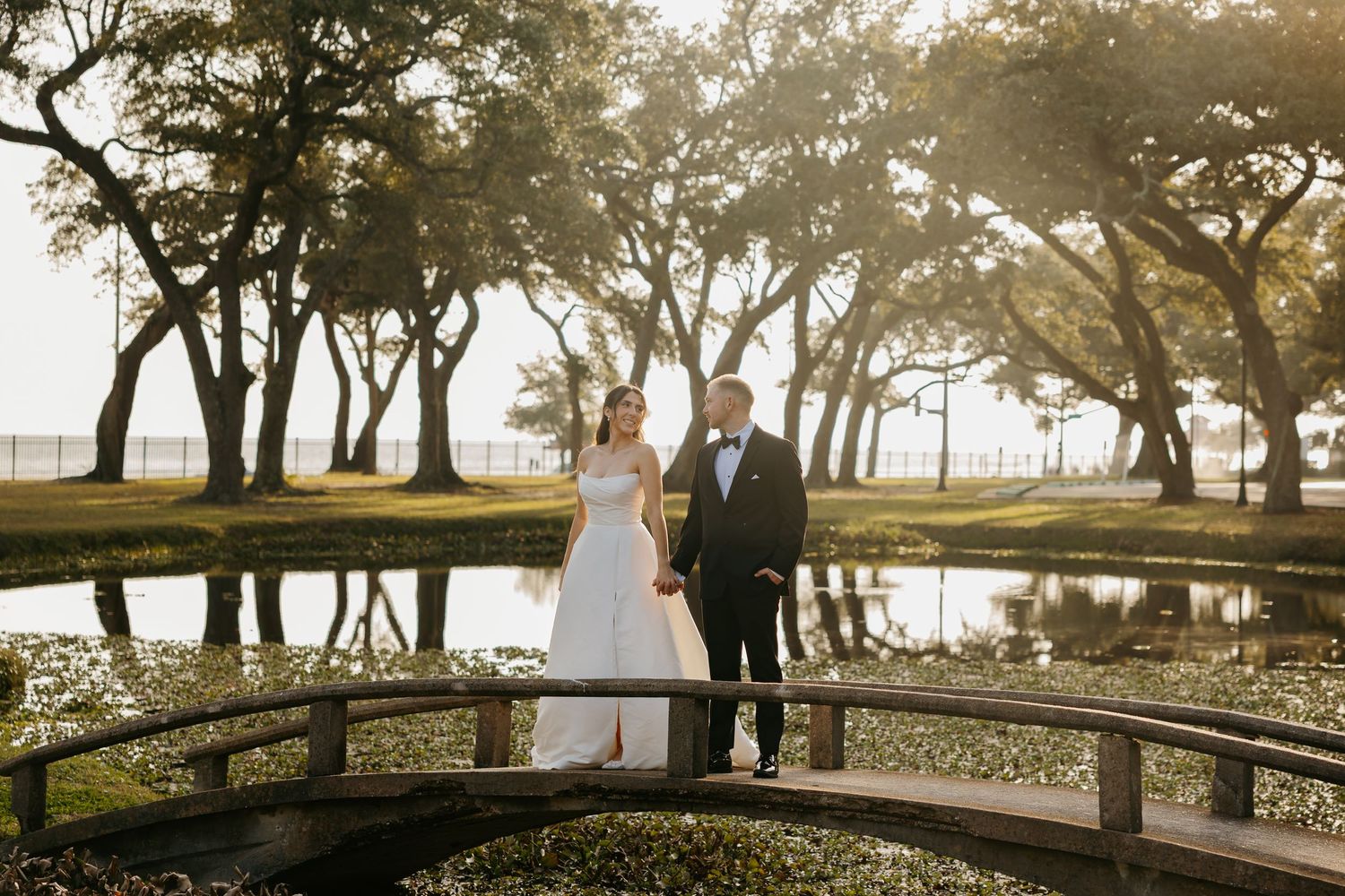 Bride and groom walk hand-in-hand across wooden bridge surrounded by moss-draped trees at sunset.