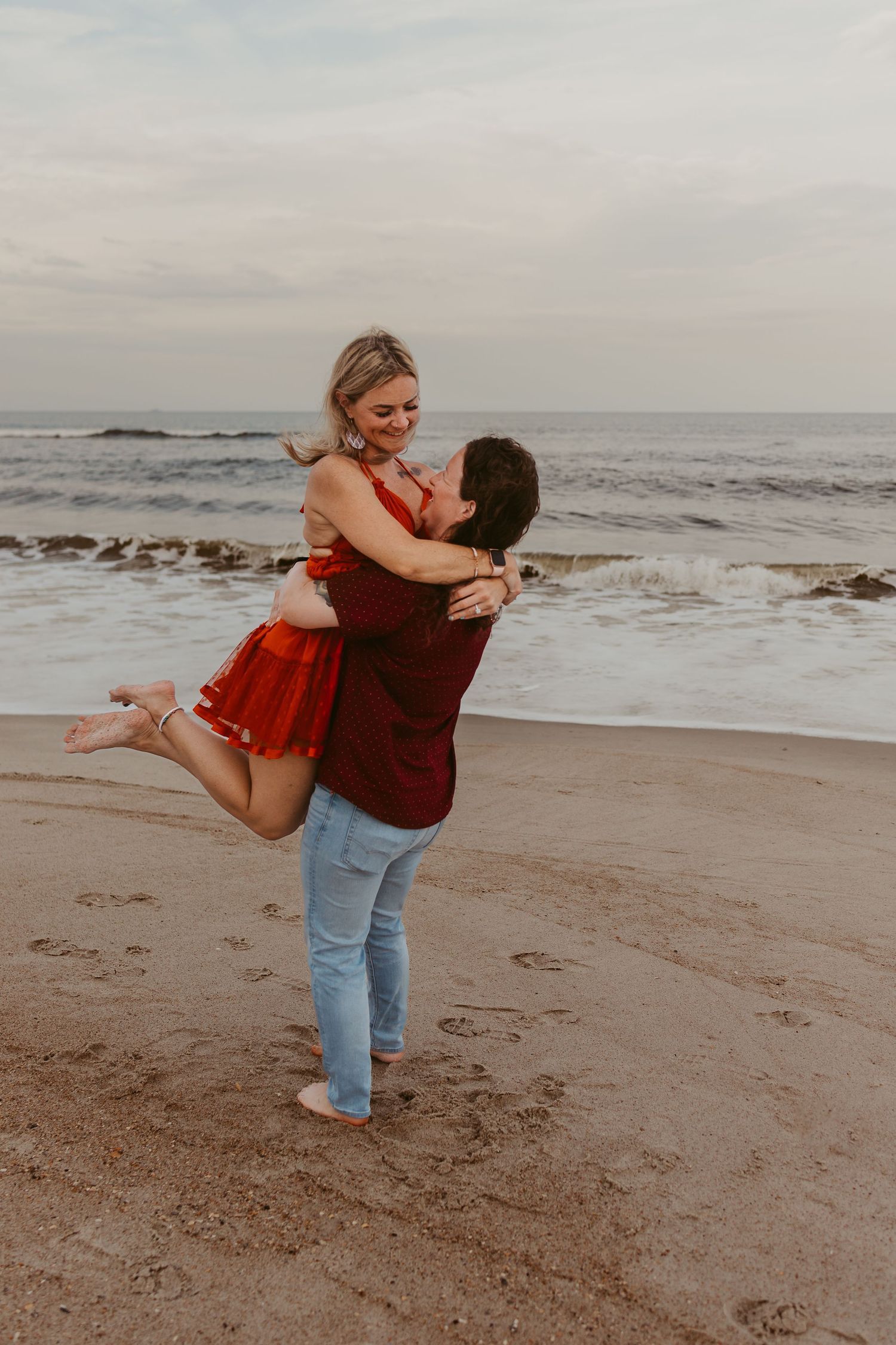 Joyful couple celebrates beach engagement as woman is lifted and spun near ocean waves.