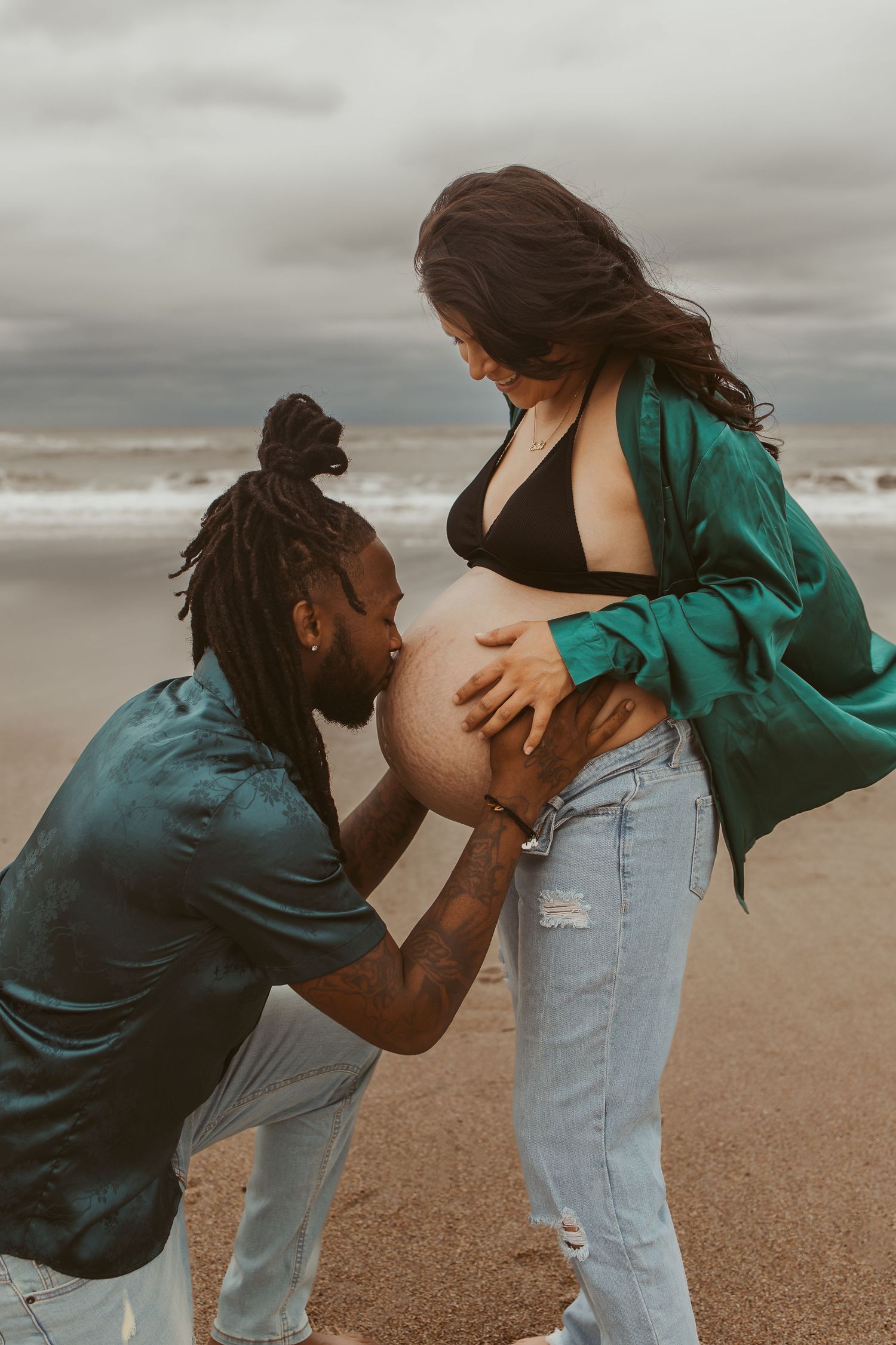 Man kneeling to kiss pregnant partners bare baby bump on sandy beach during maternity shoot.