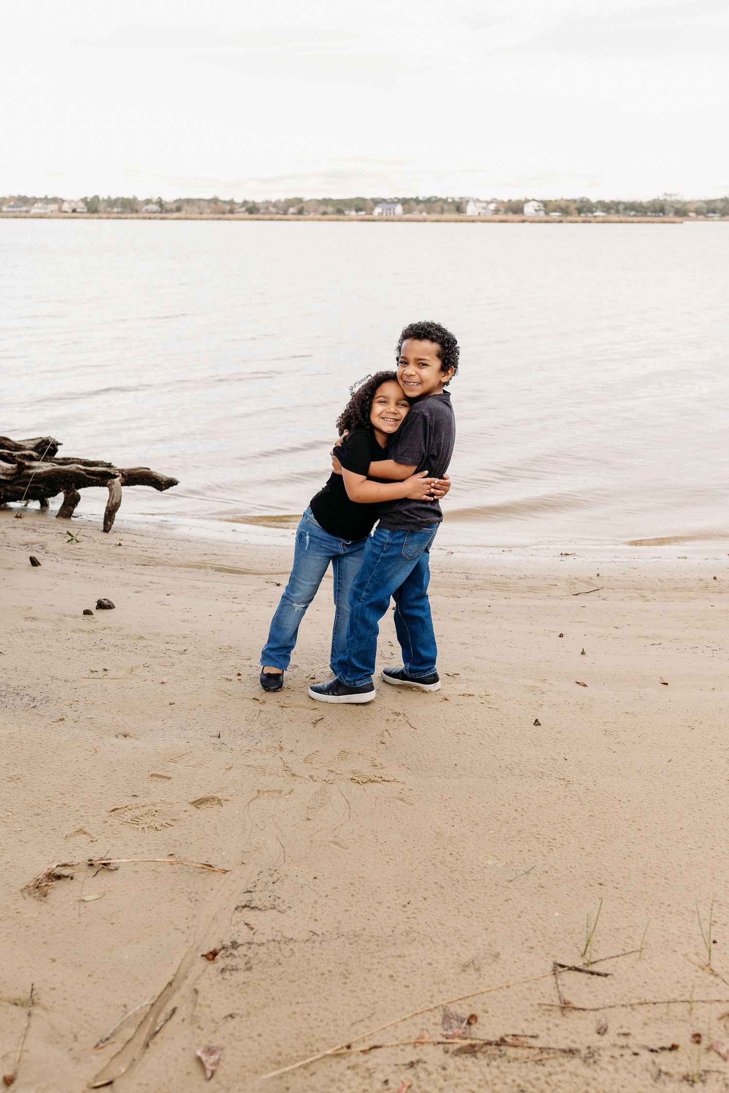 Two children hugging and smiling on a sandy beach with calm water in the background.