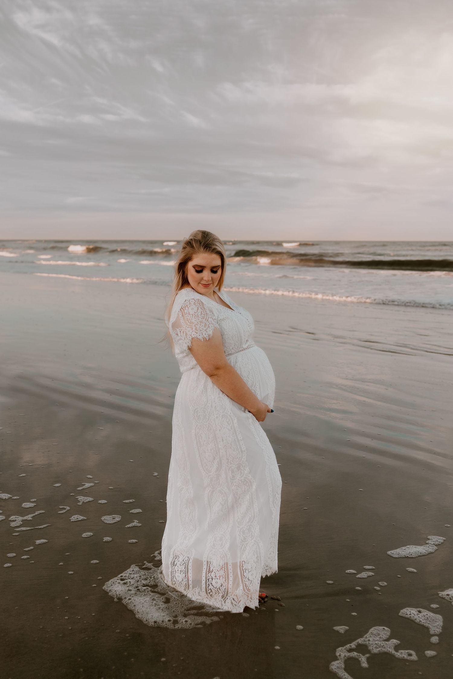 Pregnant woman in white lace dress looking down at belly while standing in ocean waves at sunset.