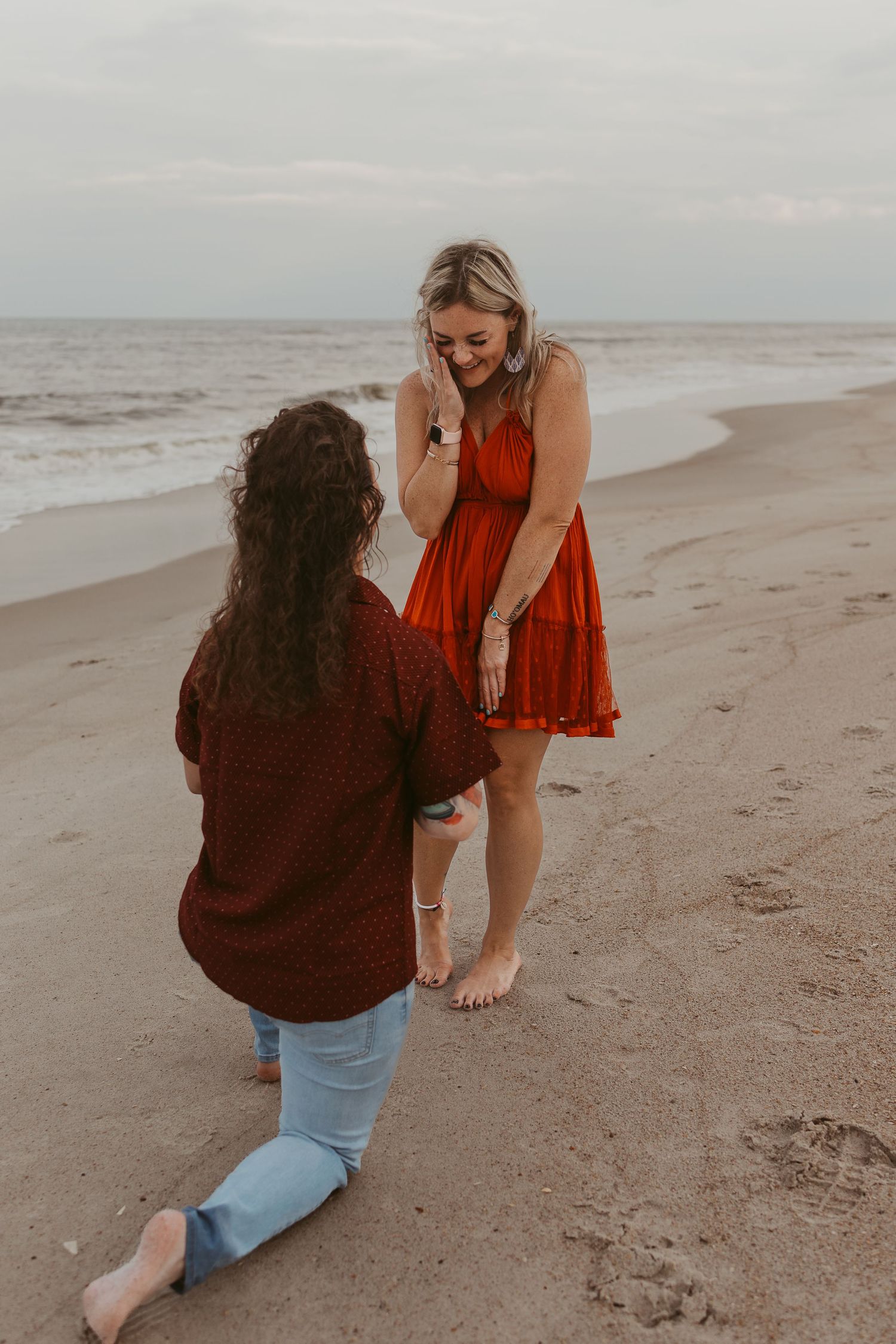 Two women share a beach proposal moment, one kneeling in the sand as the other reacts with joy.