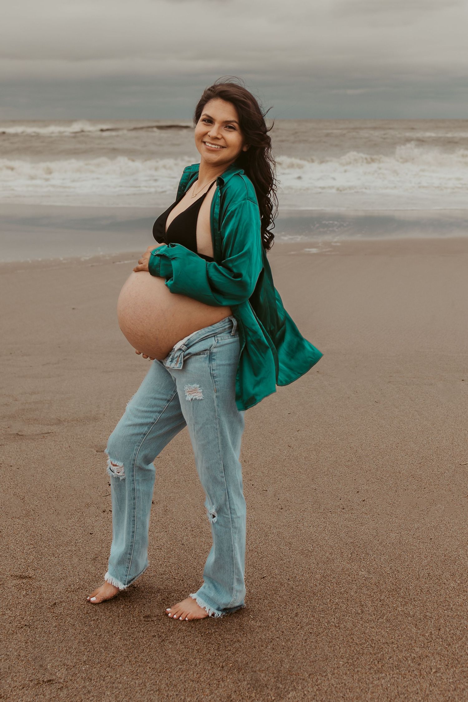 Smiling pregnant woman in teal shirt and distressed jeans posing on beach with ocean backdrop.