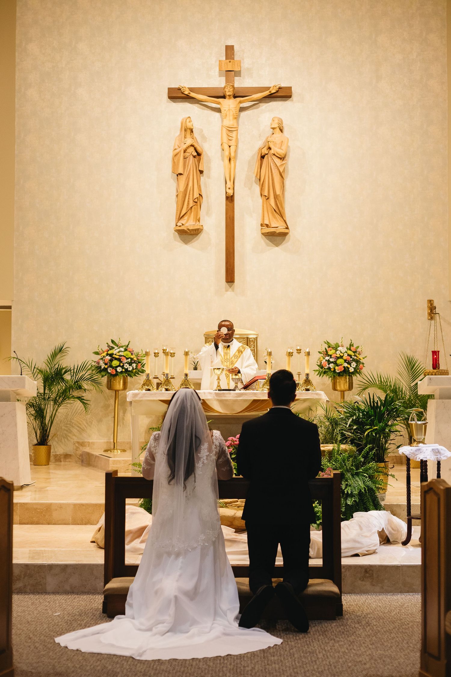 Bride and groom kneeling at altar during Catholic wedding ceremony beneath a crucifix in a warmly lit church.