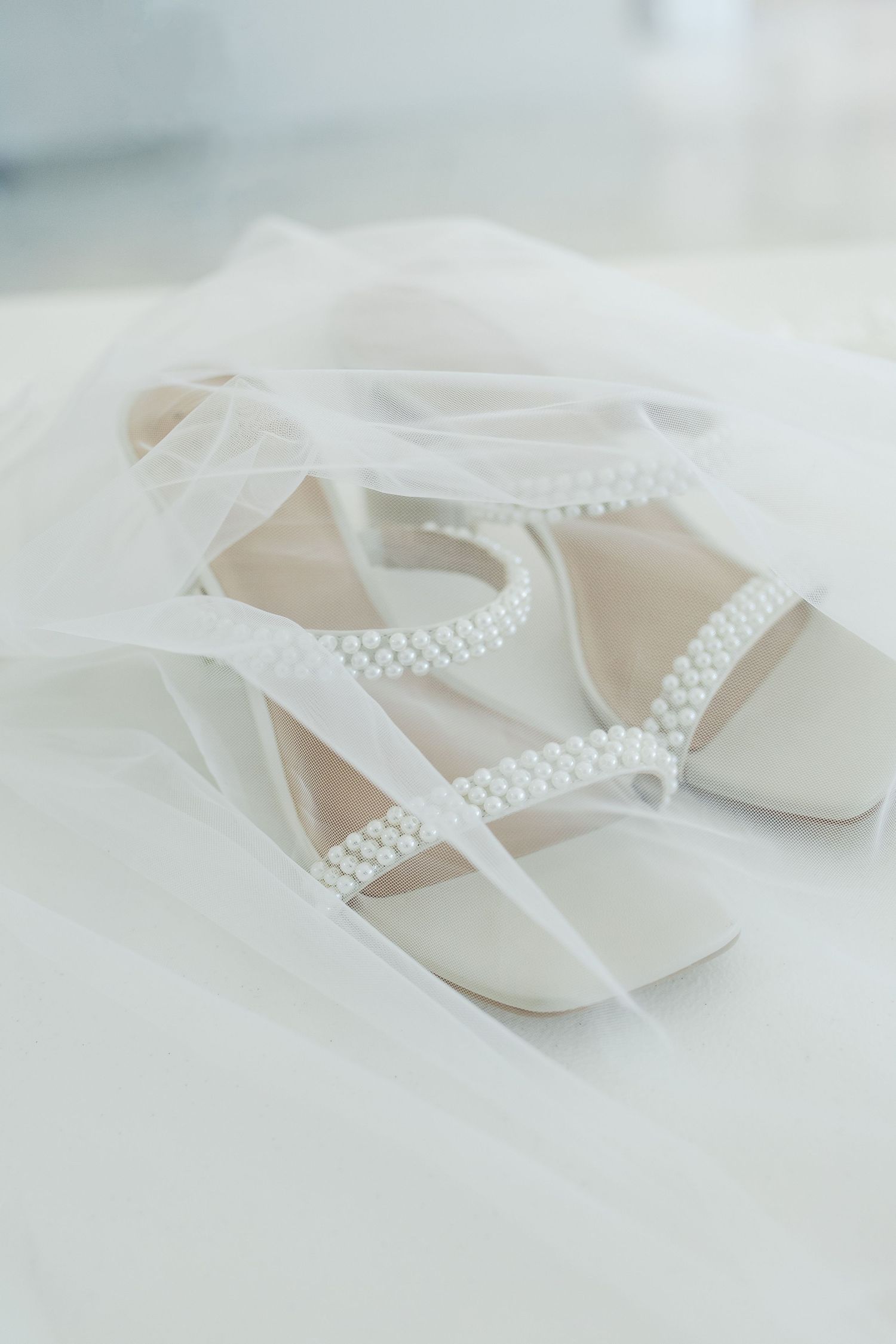 Pearl-embellished white bridal sandals resting on sheer wedding veil.