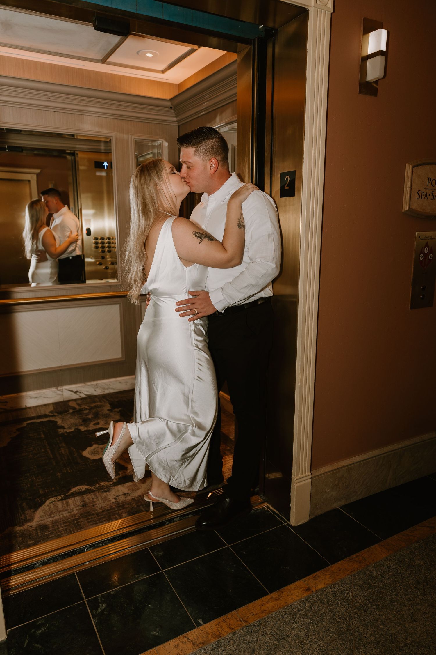 Couple sharing passionate kiss in elevator doorway, woman in white satin dress lifting heel romantically.