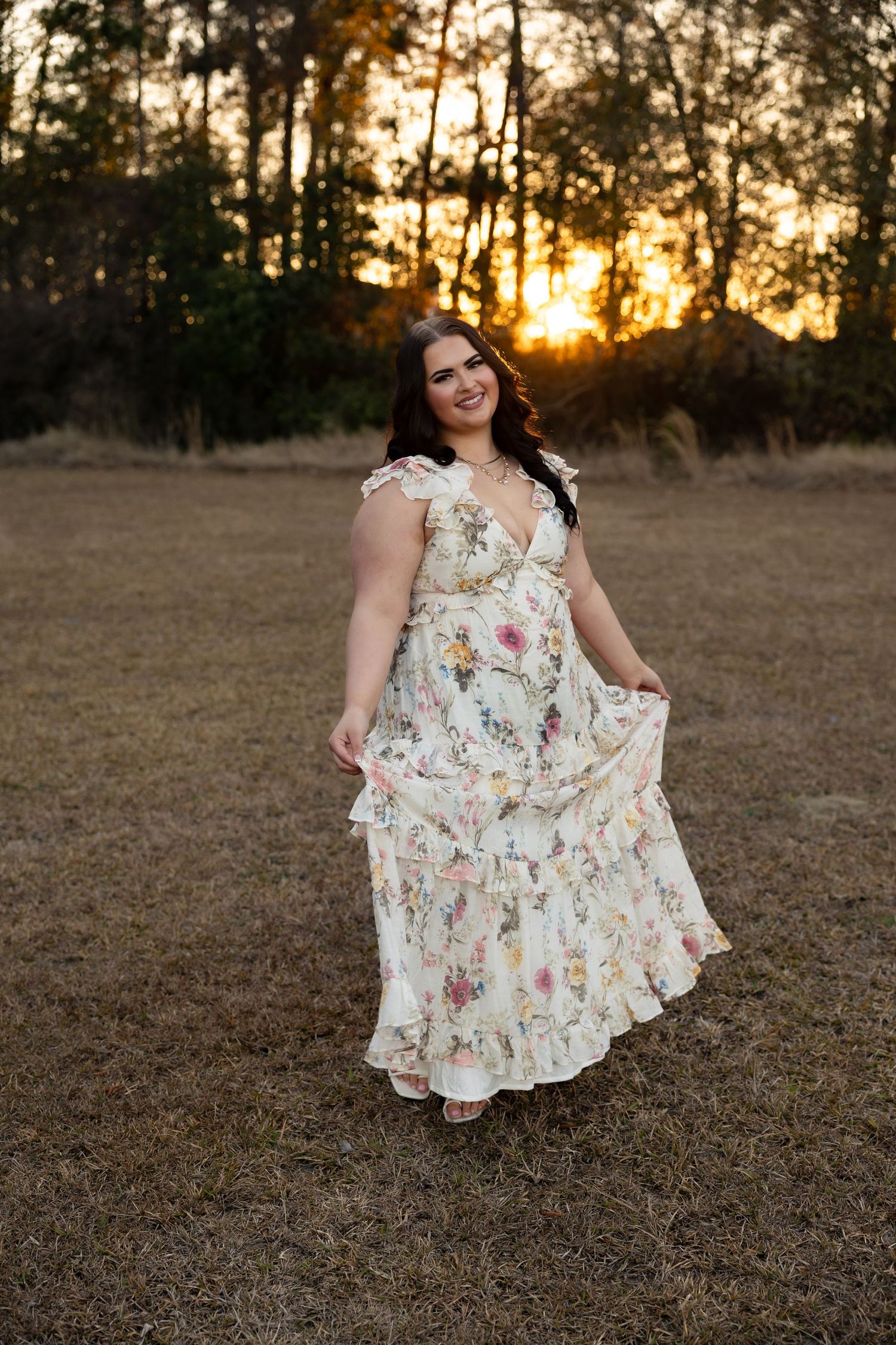 Young woman in floral maxi dress twirling at golden hour sunset in open field.