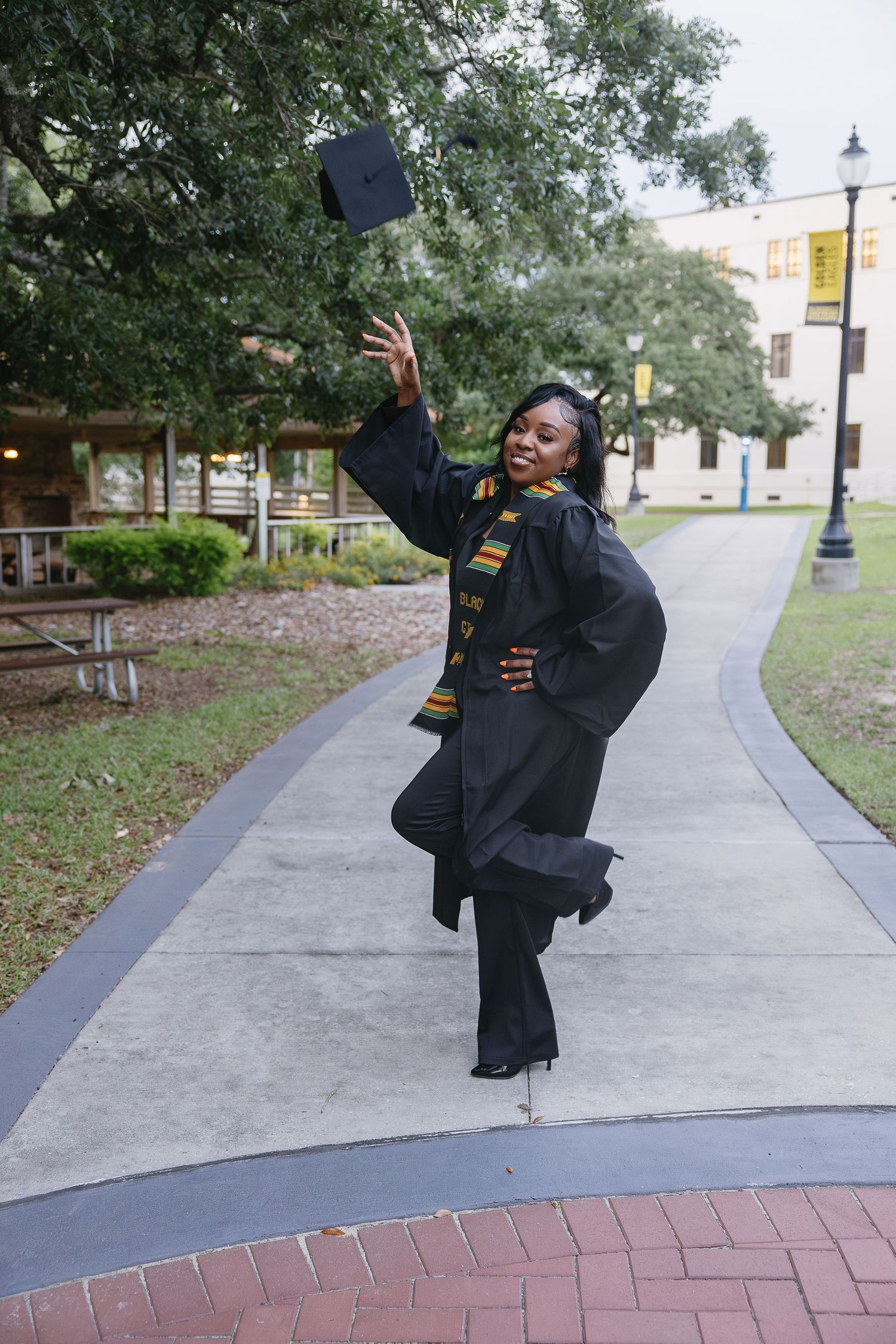 A joyful graduate in cap and gown tosses her cap while walking on a campus pathway.