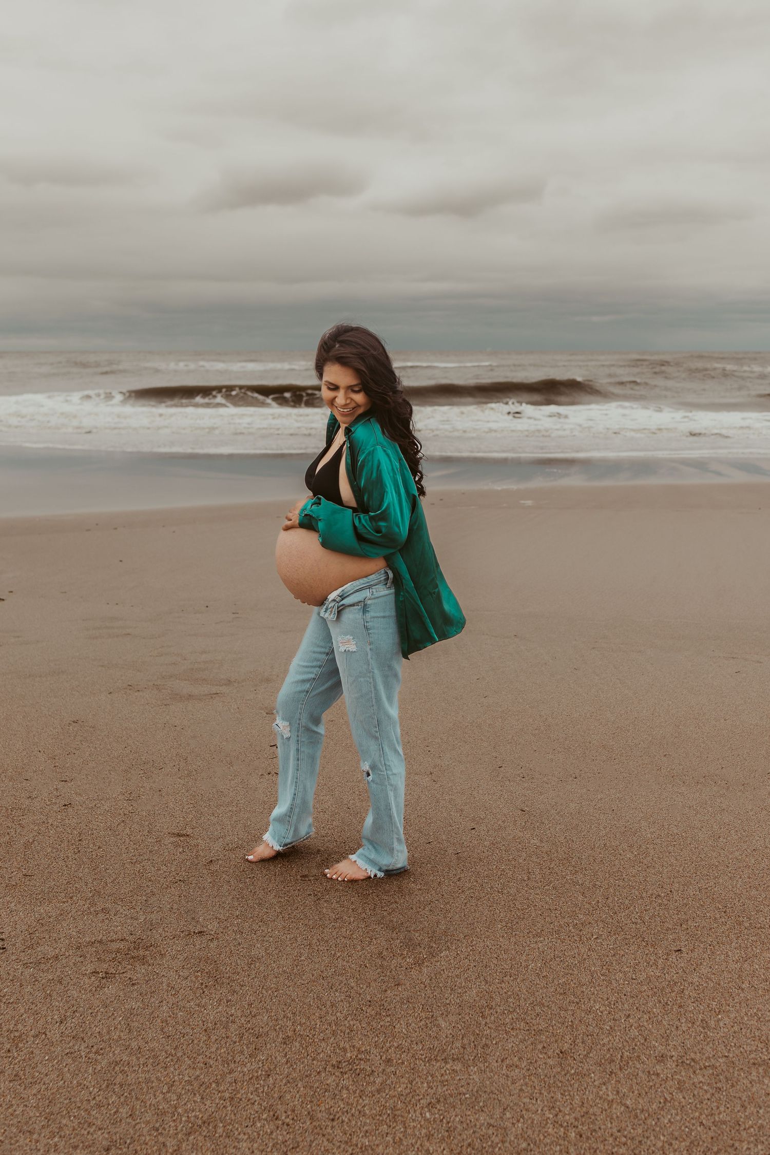 Pregnant woman in green shirt and ripped jeans cradling baby bump on sandy beach with ocean waves.