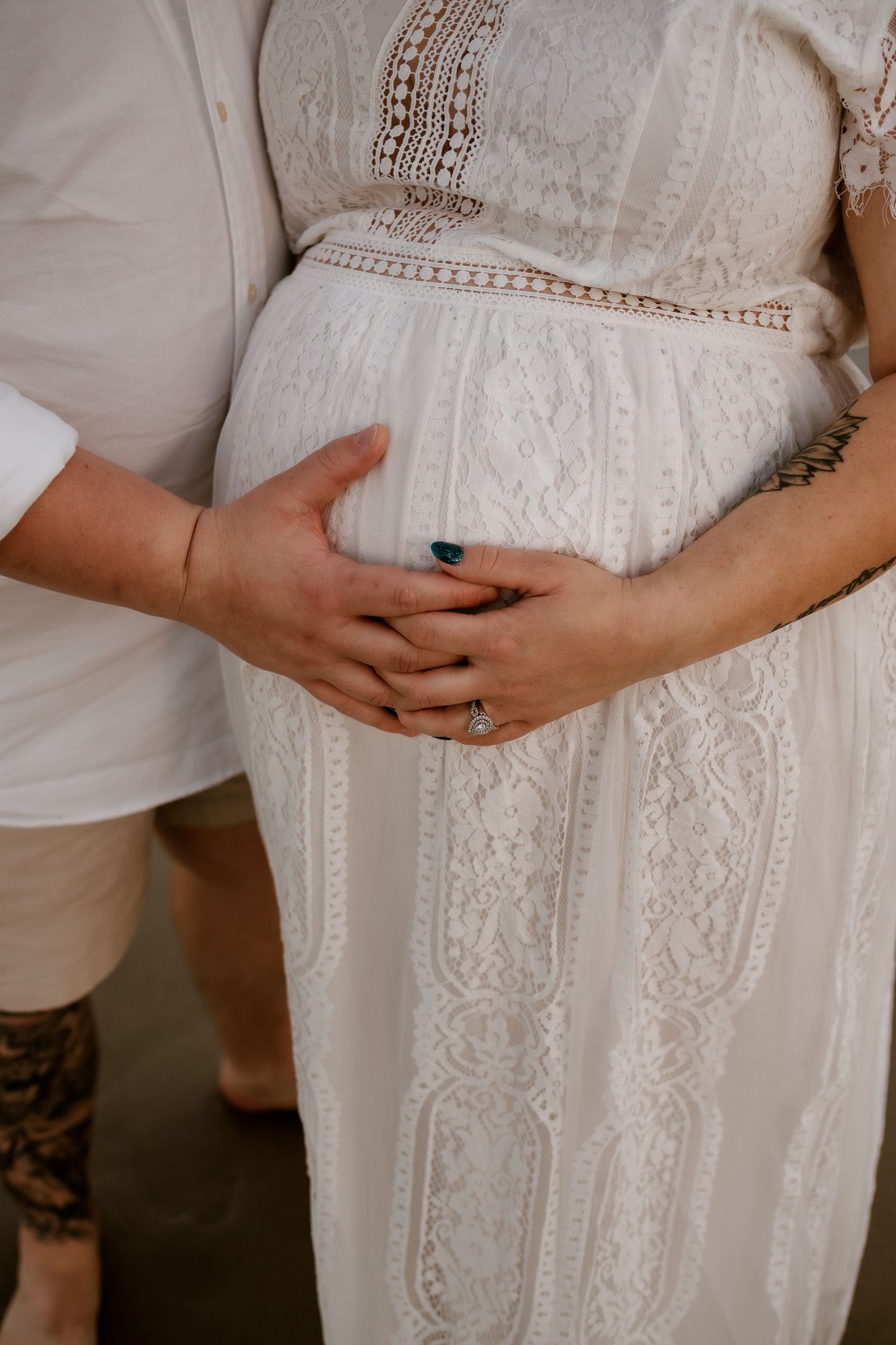 Close-up of couple's hands cradling pregnant belly in white lace maternity dress, diamond ring visible.