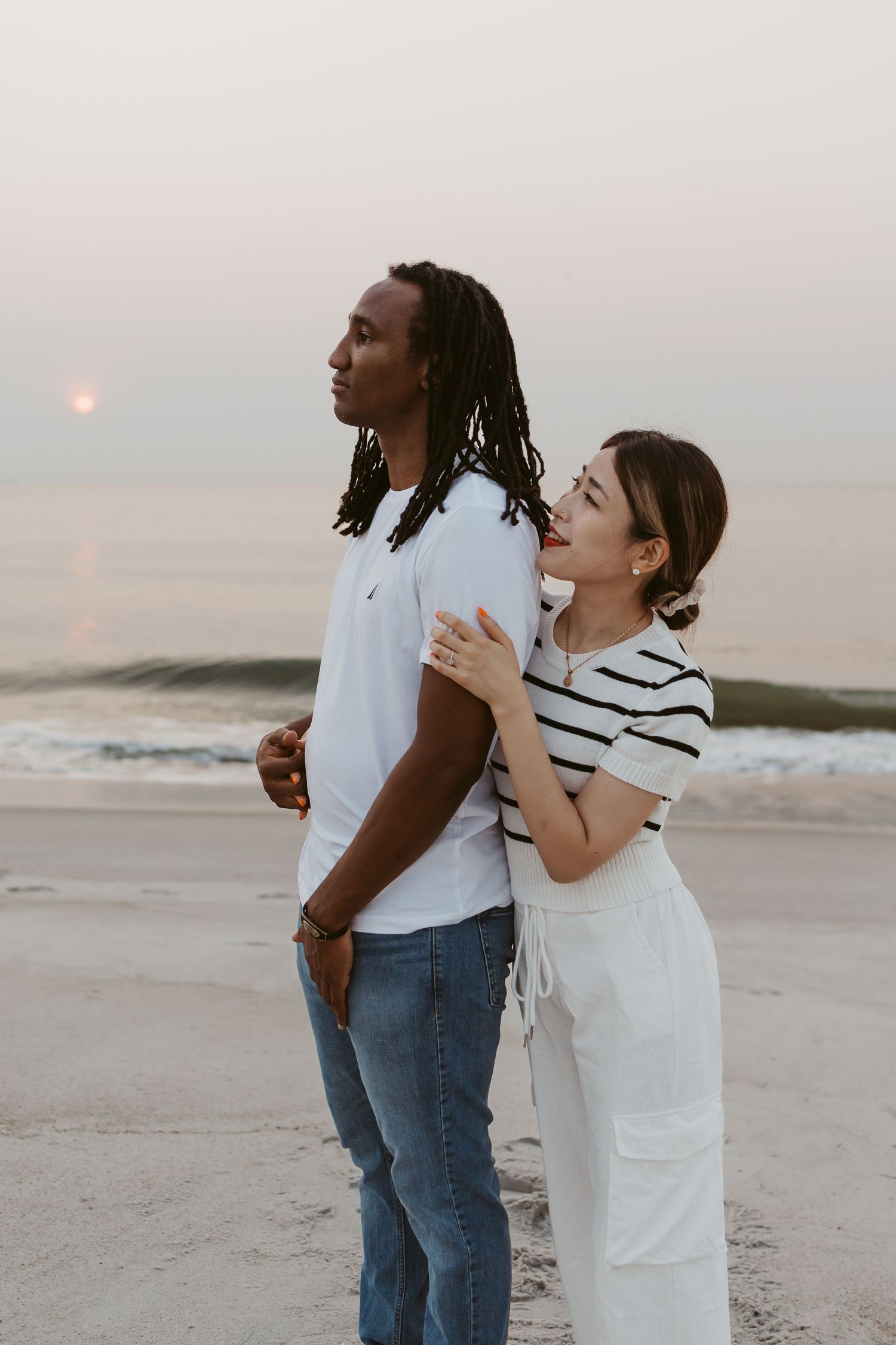 Woman in striped shirt hugging man from behind on a beach at sunset.