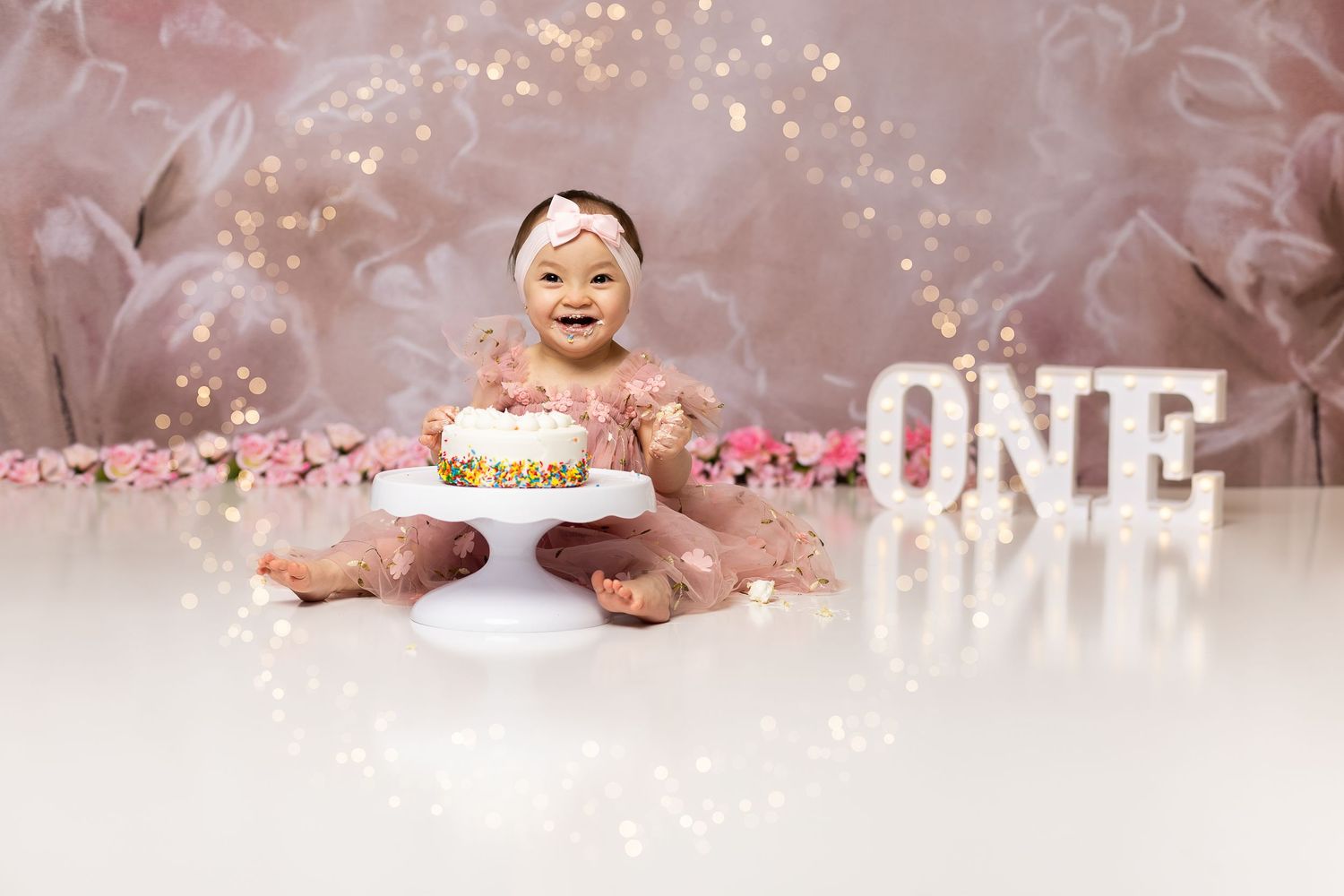 A baby in a pink tutu smiles beside a cake during a first birthday cake smash session.