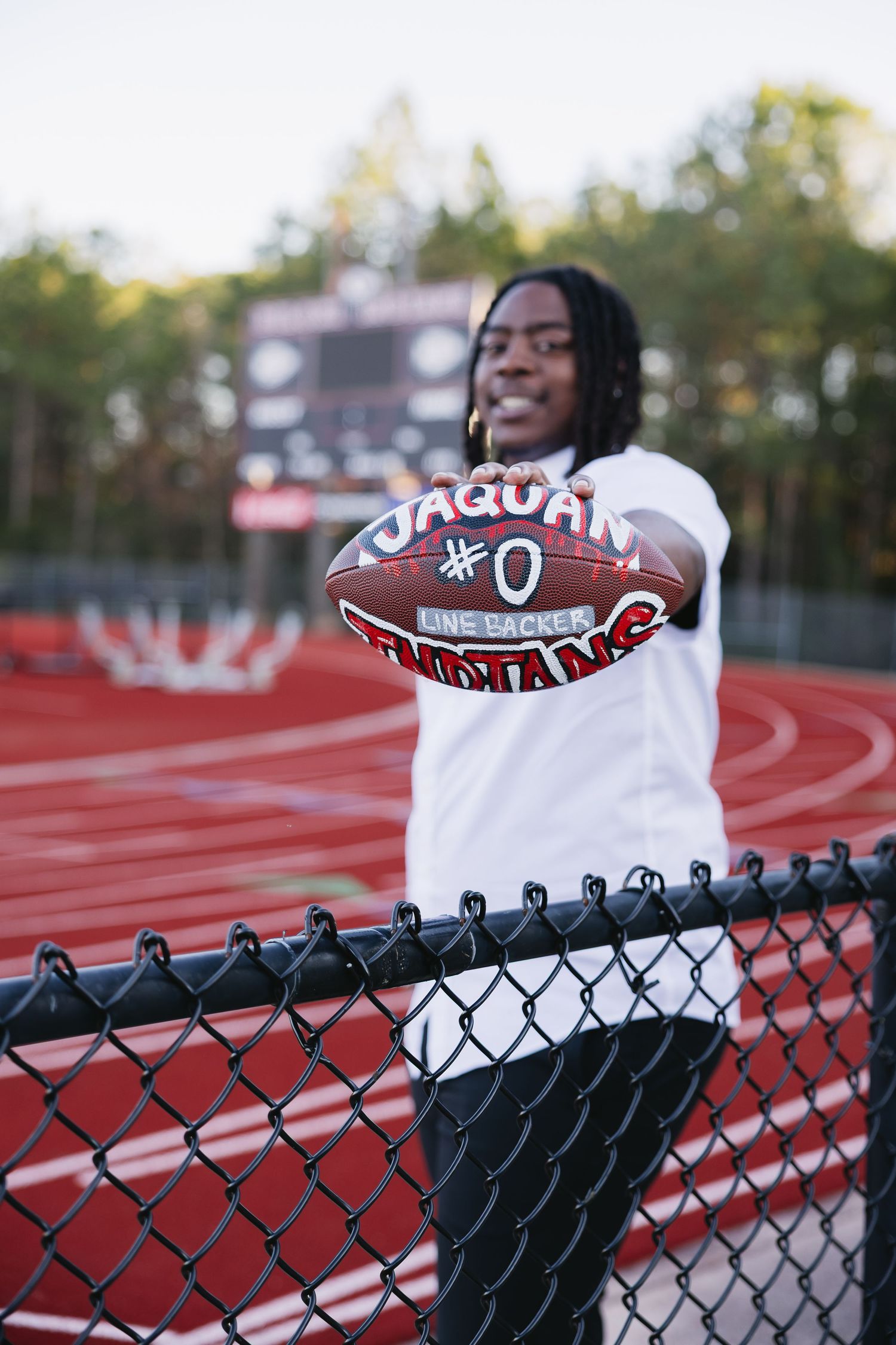 Senior football player holding custom Jaquan #0 Indians football over chain-link fence at stadium.