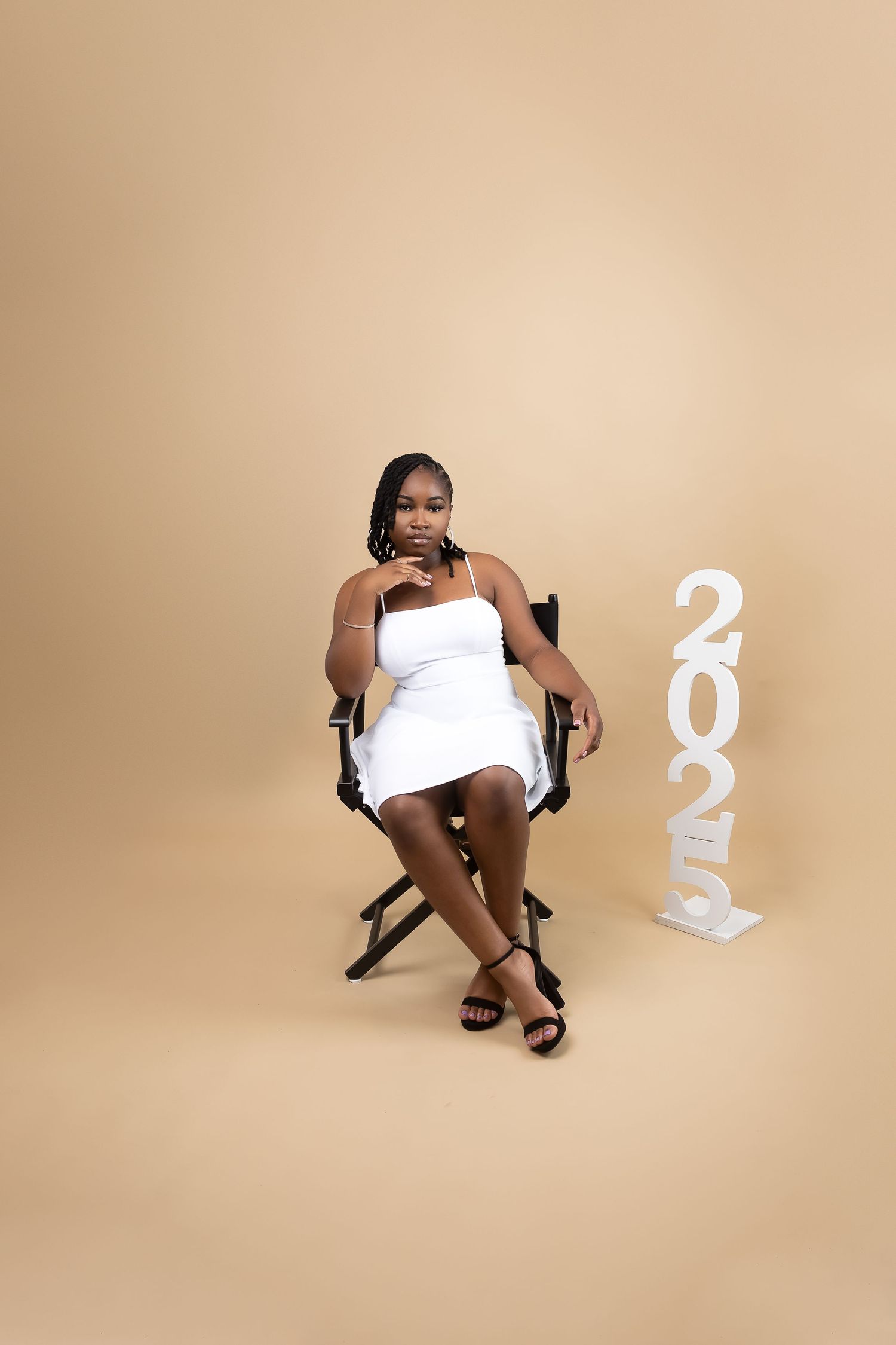 Young woman in white dress seated confidently in chair beside 2025 sign on beige backdrop.