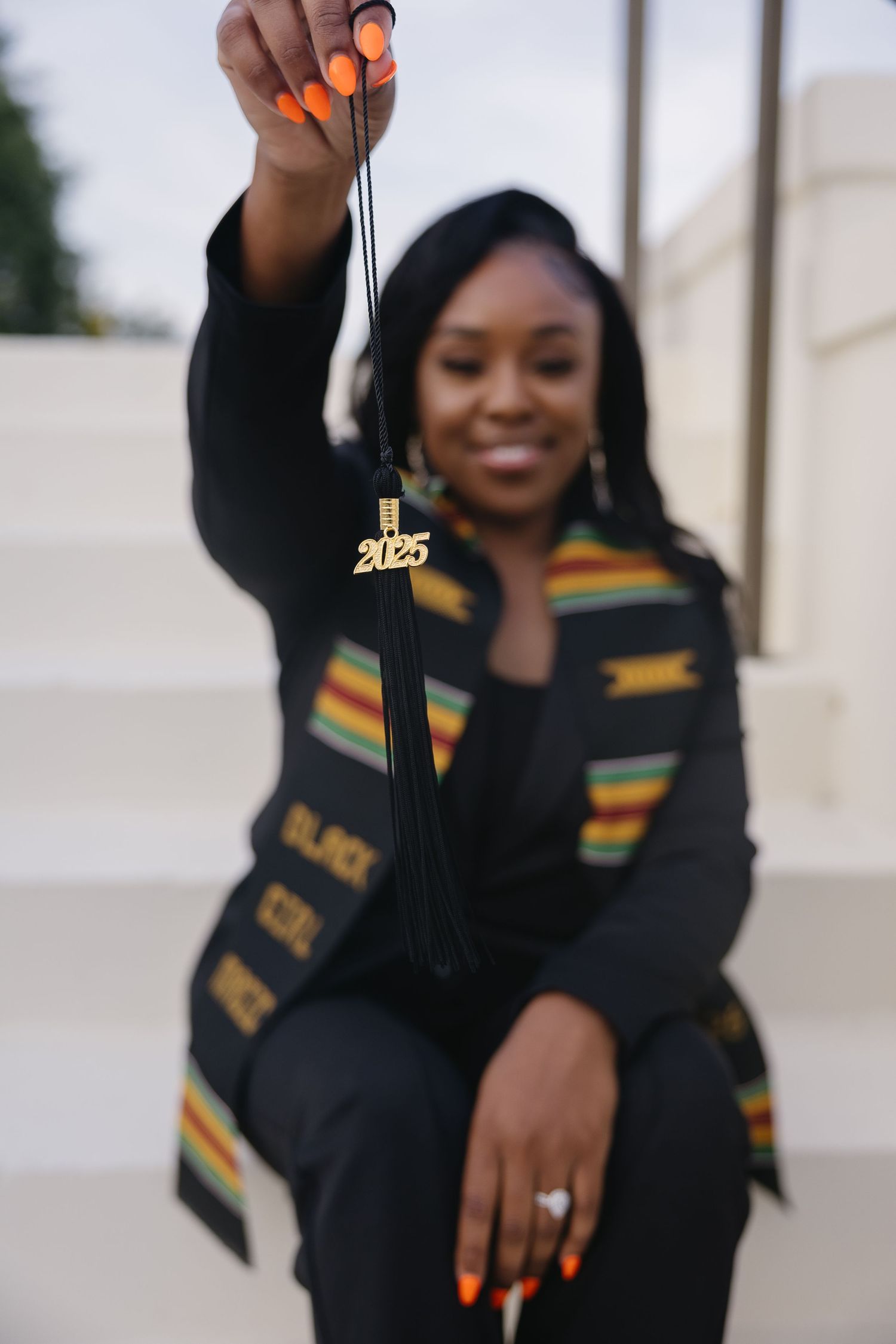 A graduate holds up a gold 2025 tassel charm while wearing a Black Girl Magic kente stole.