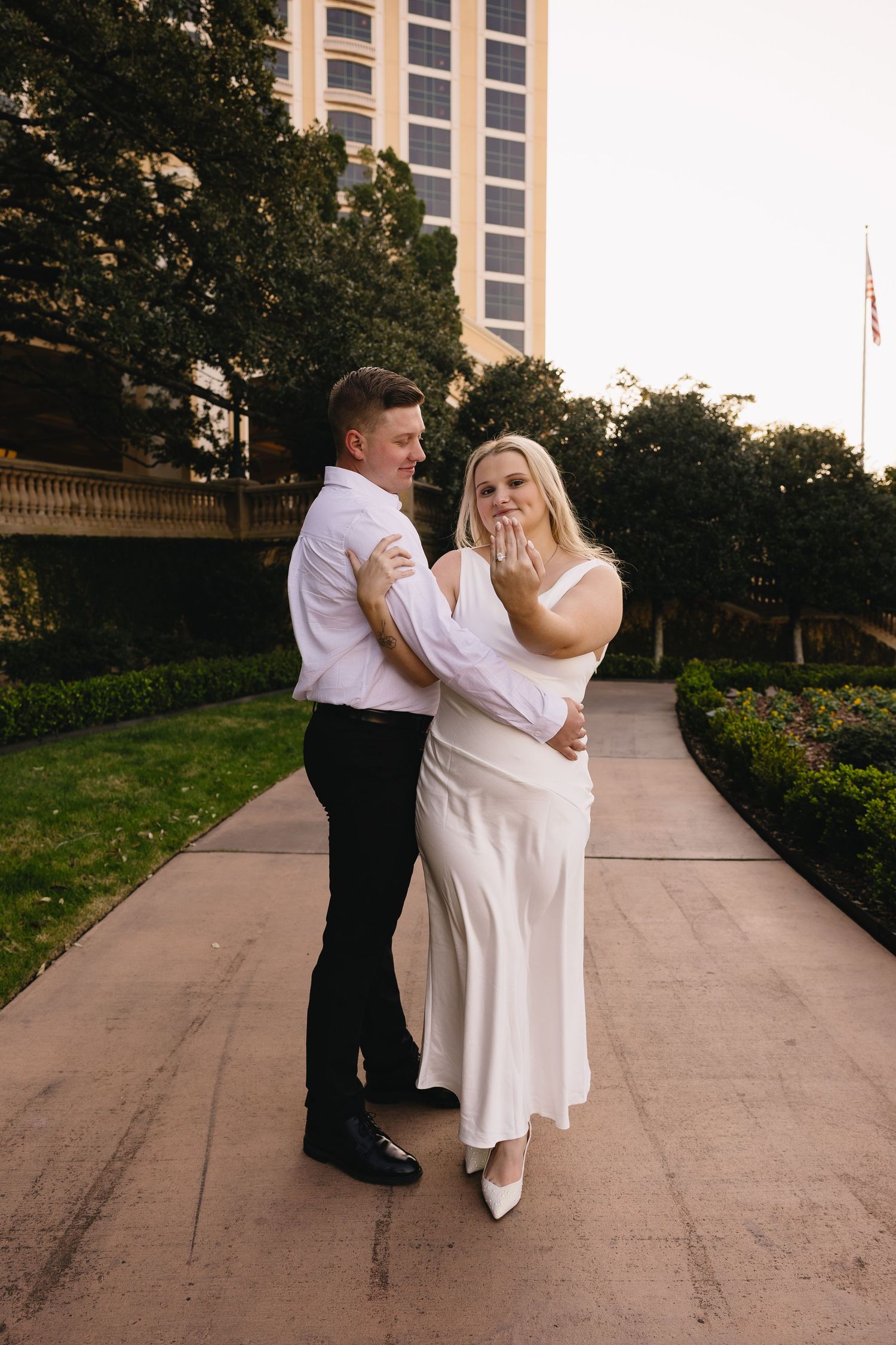 Joyful woman in white dress shows engagement ring while embracing man in white shirt on garden pathway.