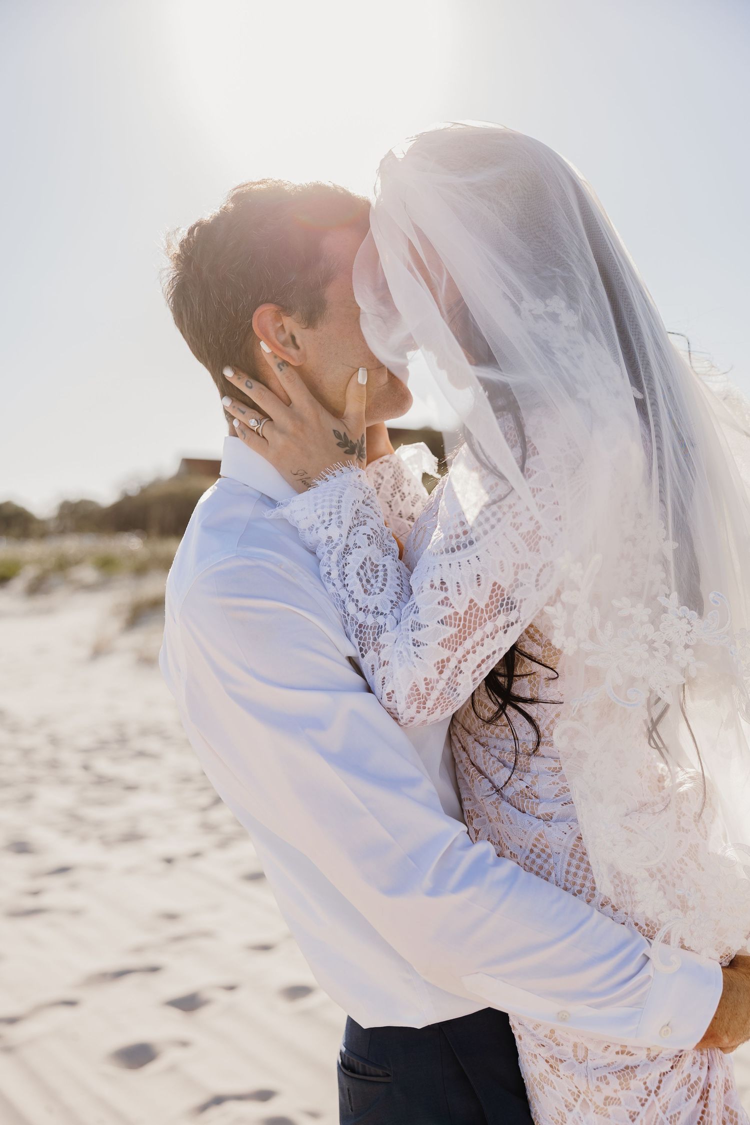 Newlyweds sharing a romantic kiss on a sunny beach, bride in lace dress and flowing veil.