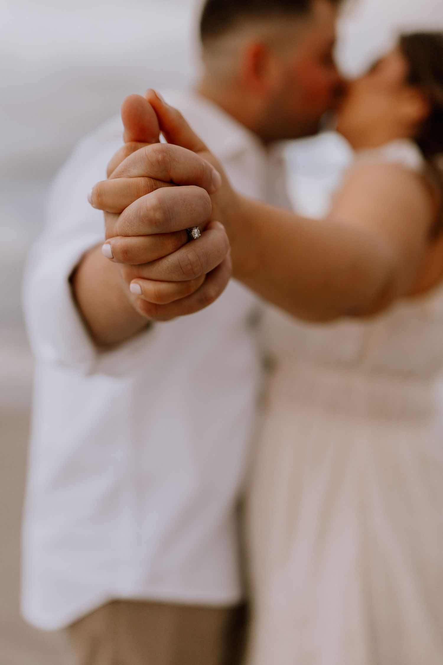 Engaged couple kissing on a beach while holding hands, with a diamond ring visible in the foreground.