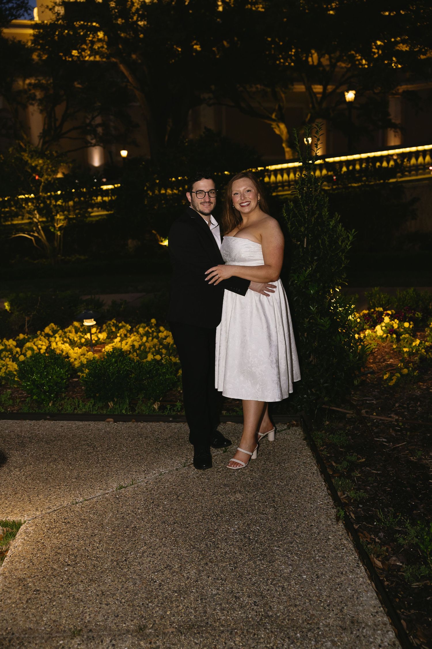 Newlyweds embracing on garden pathway at night surrounded by illuminated yellow flowers.