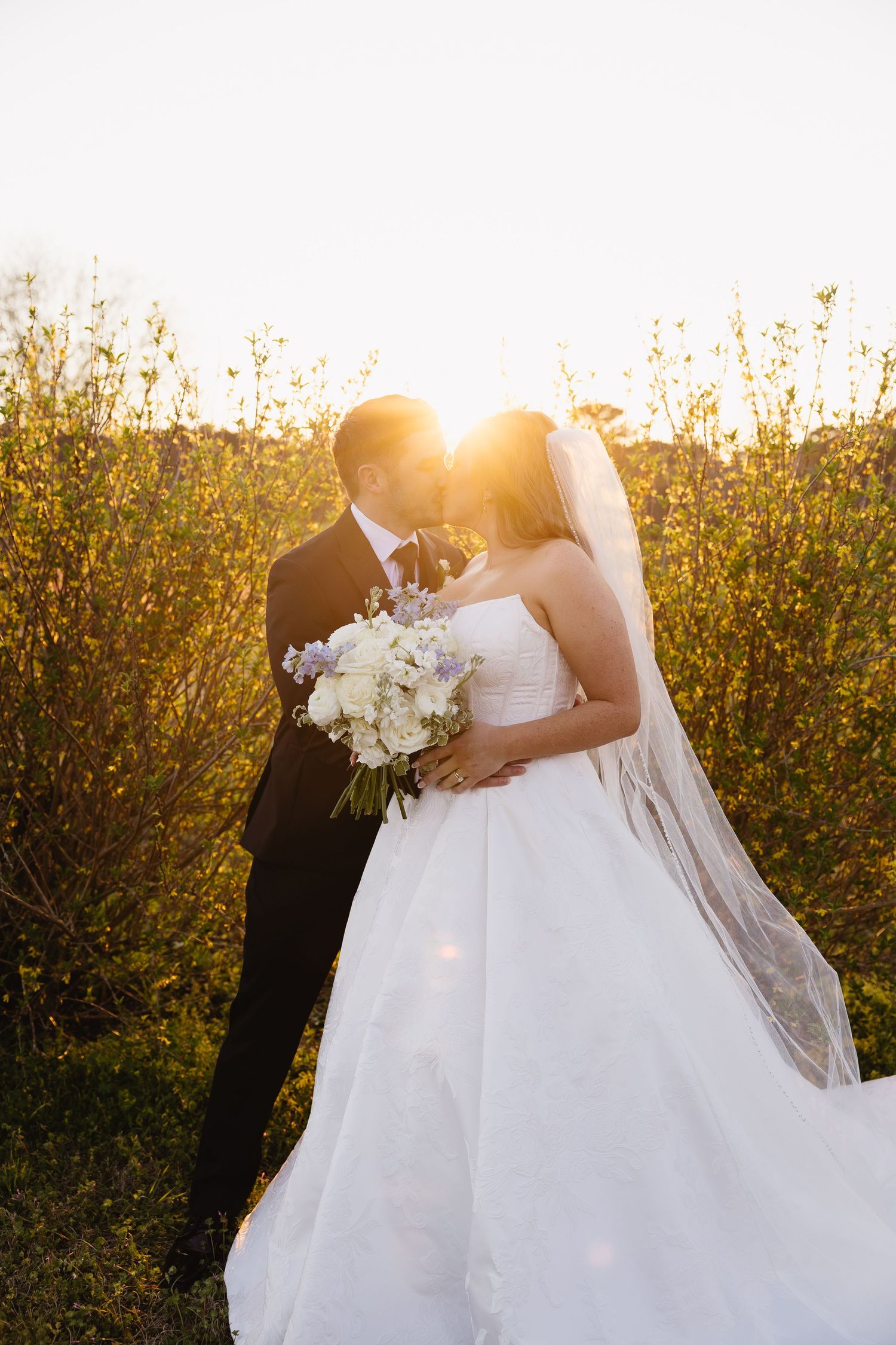 Bride and groom kissing at sunset in field with bride holding white bouquet and wearing veil.