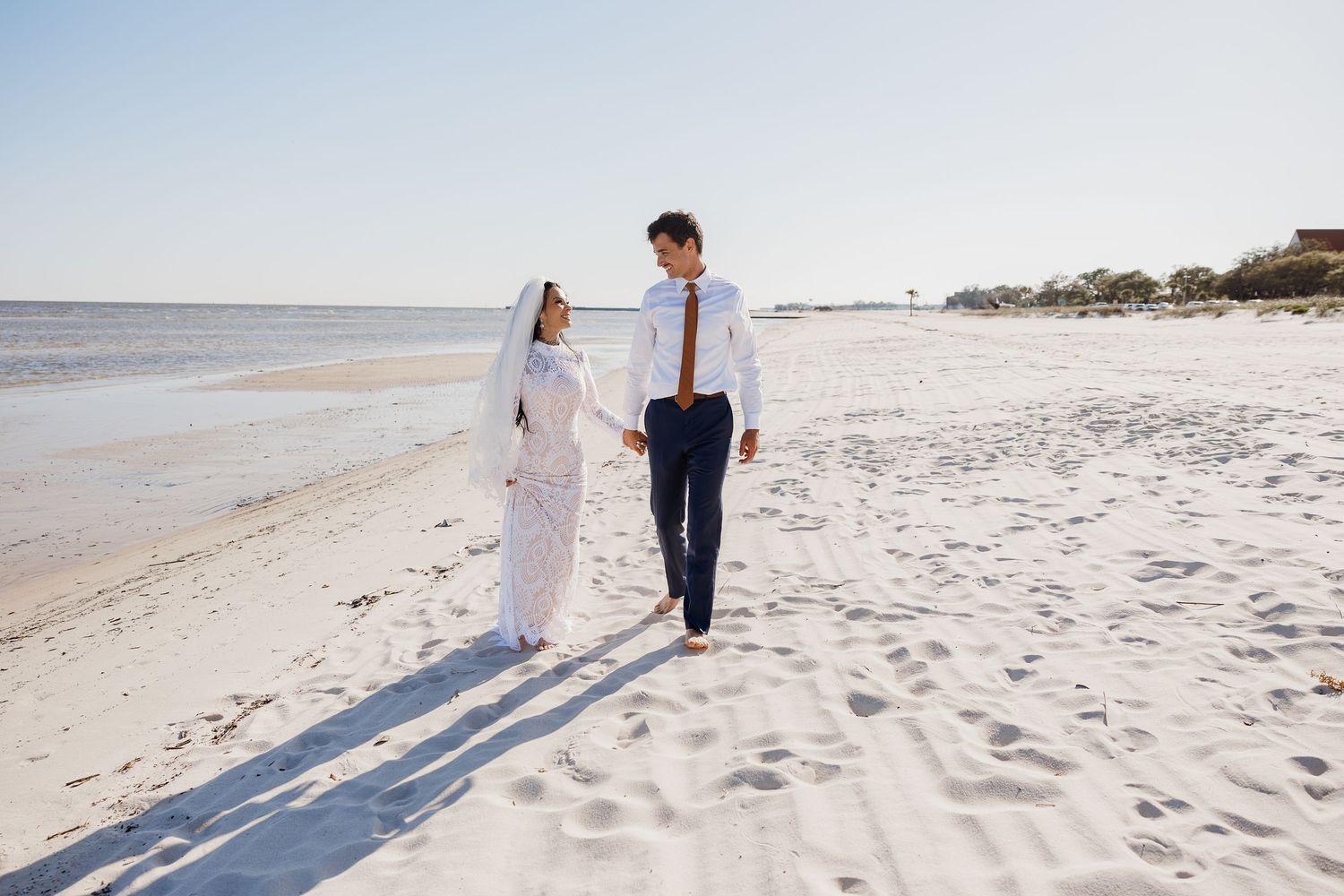 Bride and groom walk hand in hand along a sunlit white sand beach after wedding.