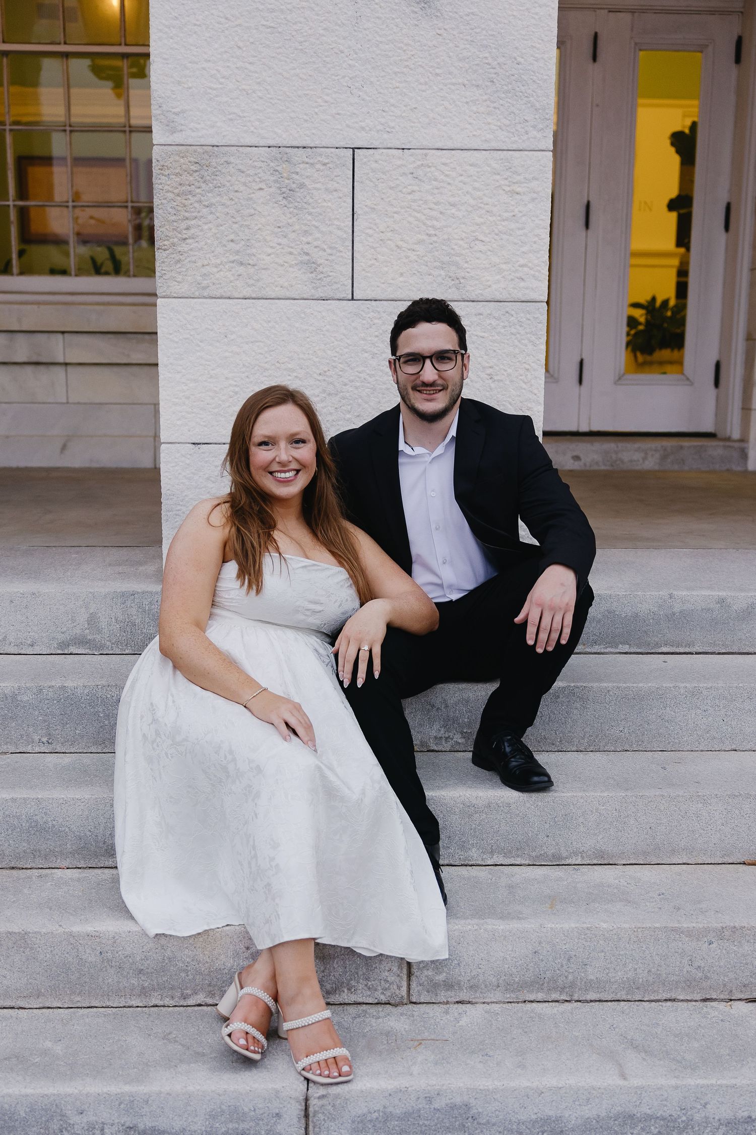 Smiling bride in white dress and groom in black suit sitting together on stone steps.