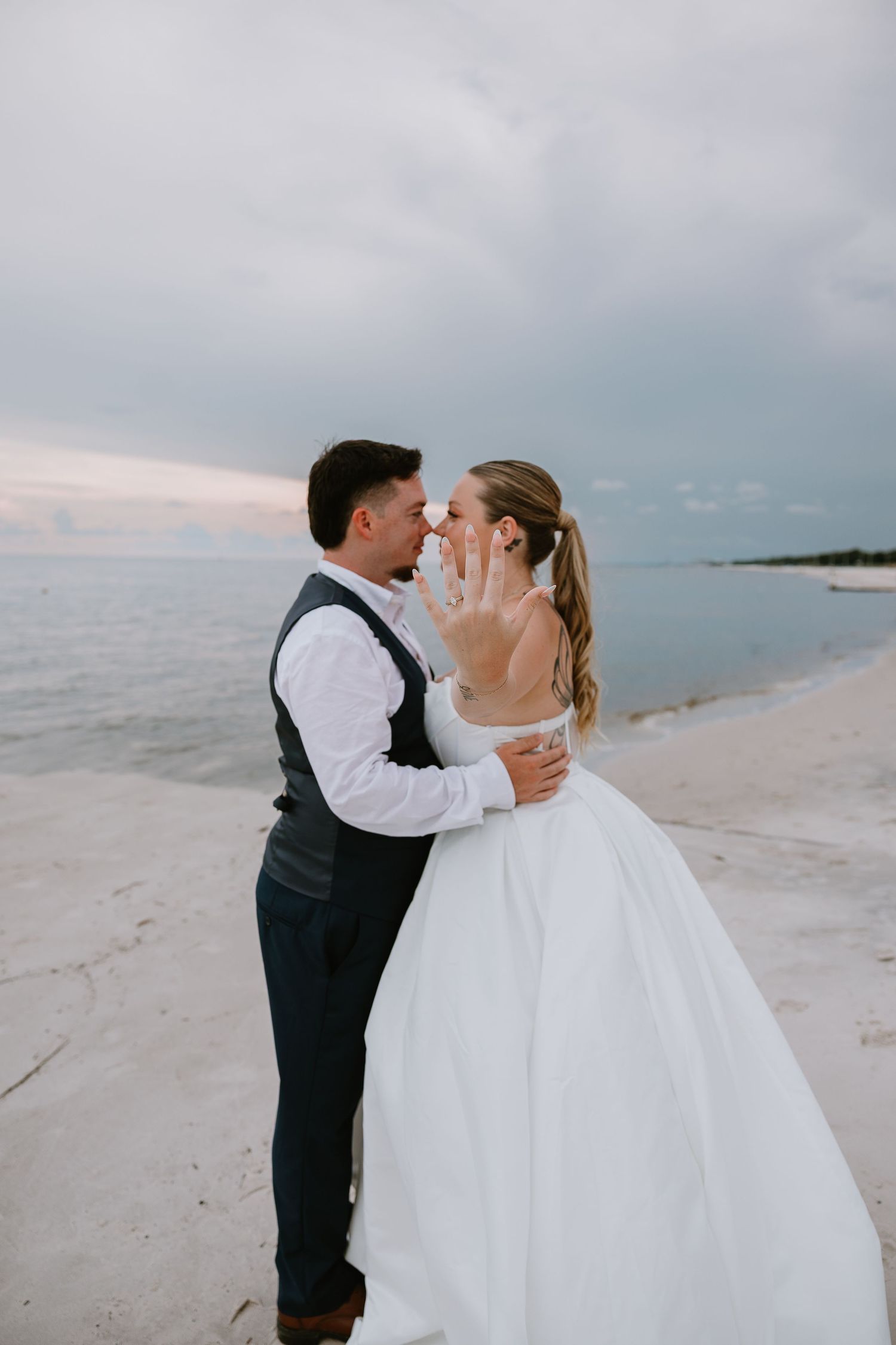 Bride showing off wedding ring while sharing a tender moment with groom on a serene beachfront.