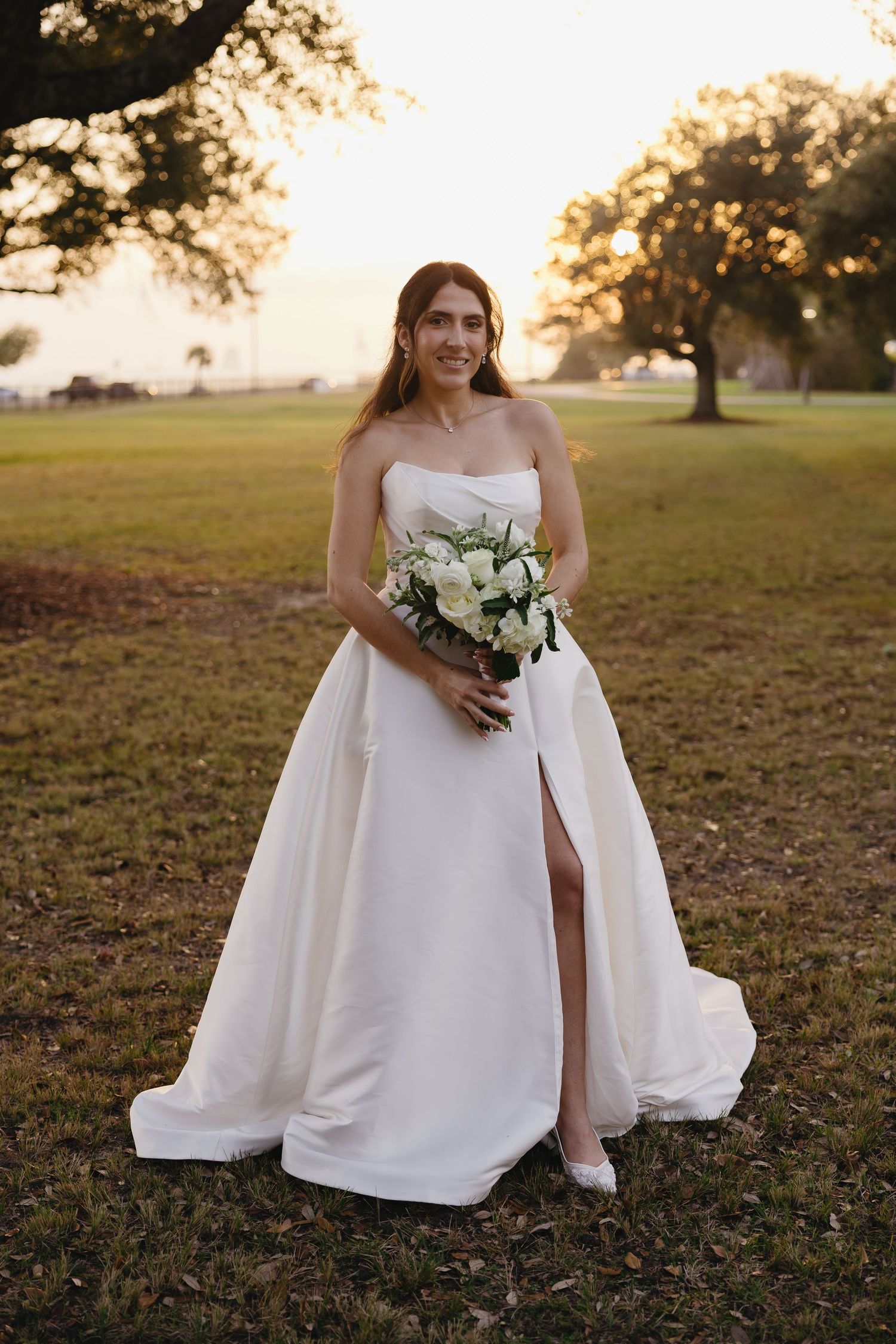 Radiant bride in strapless white ball gown holds a white bouquet at golden hour in a park.