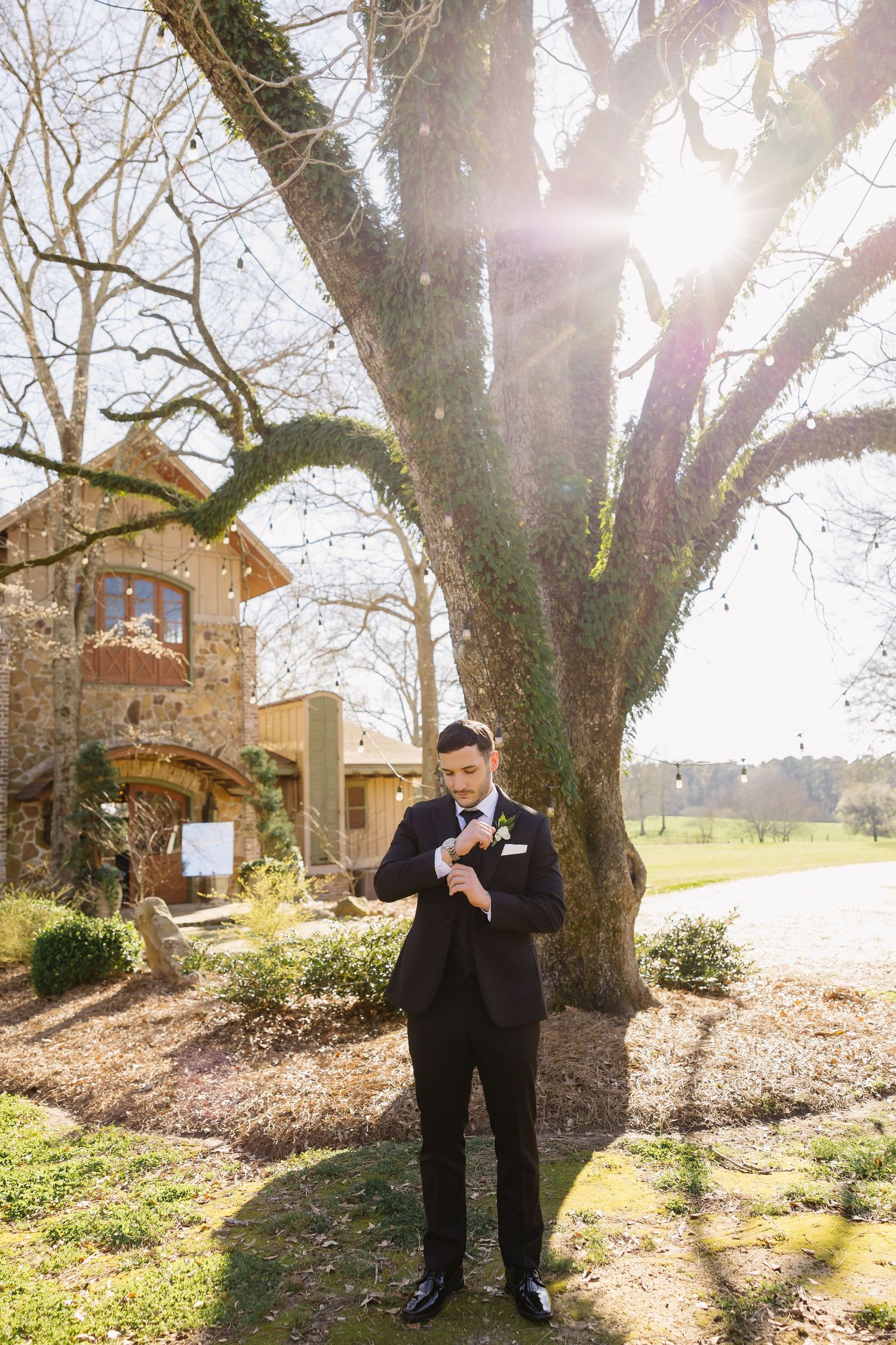 Groom in black tuxedo adjusting cufflinks outdoors near large tree with sunlight streaming through branches.