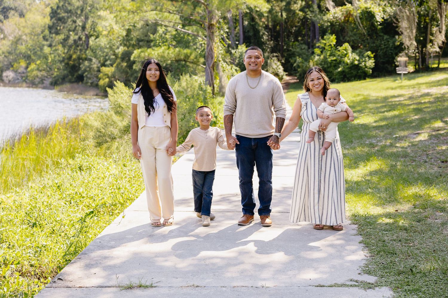 Smiling family of five walking hand-in-hand along a sunny park pathway.