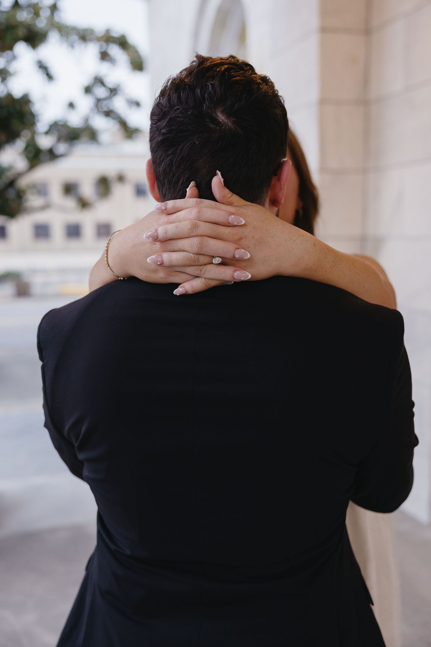 Couple embracing tenderly, woman's hands with engagement ring clasped around man's neck in romantic pose.