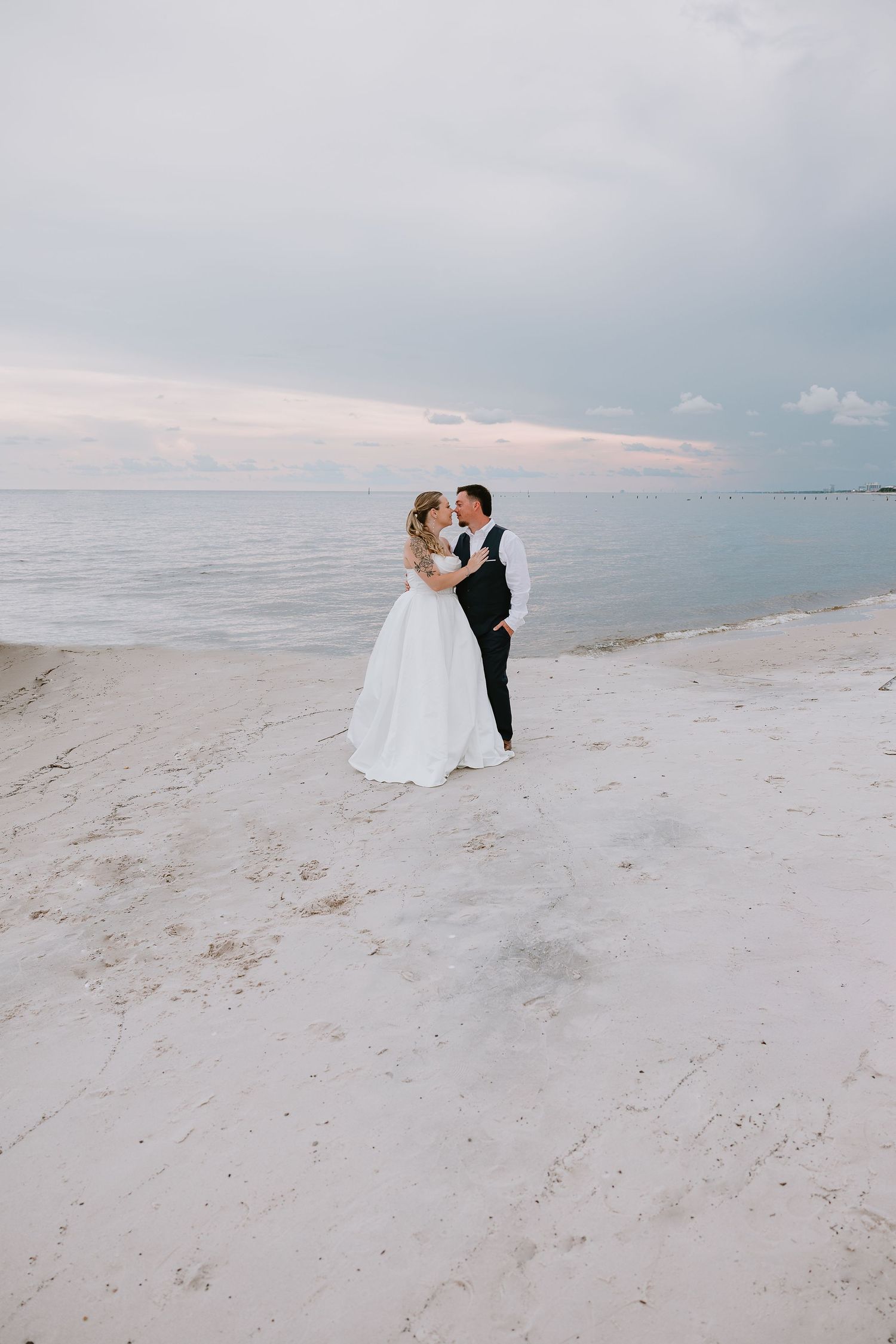 Bride and groom sharing a romantic kiss on a white sand beach with pastel cloudy sky at dusk.