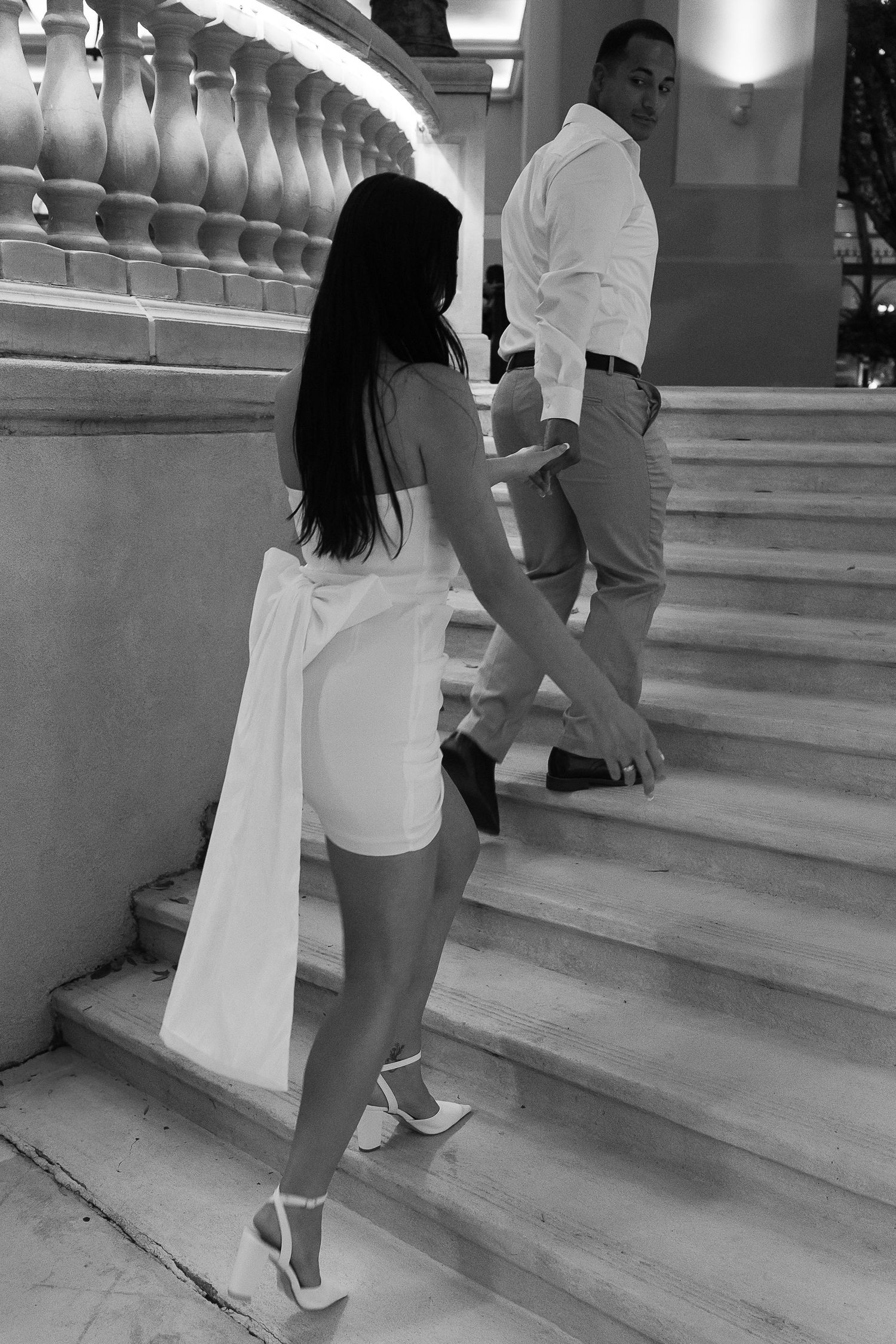 Black and white photo of couple climbing grand marble staircase, woman in white mini dress and heels holding mans hand.