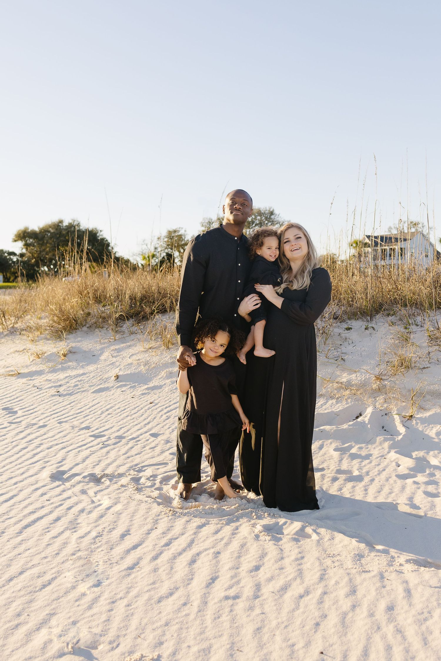 A family of four dressed in black poses together on a white sand beach at sunset.