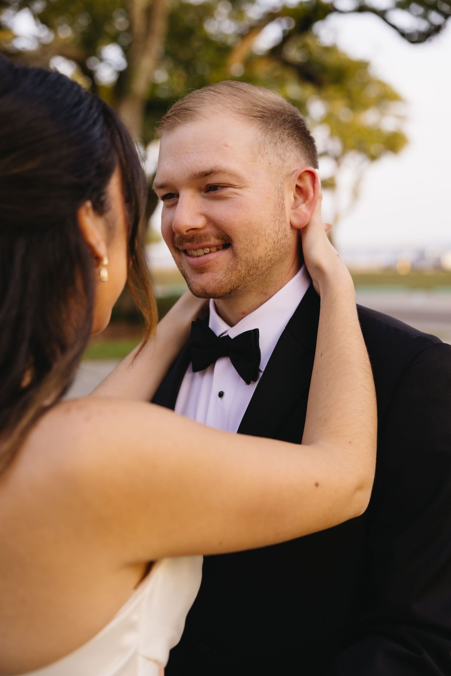Smiling groom in black tuxedo and bow tie gazes at bride who tenderly touches his face outdoors.