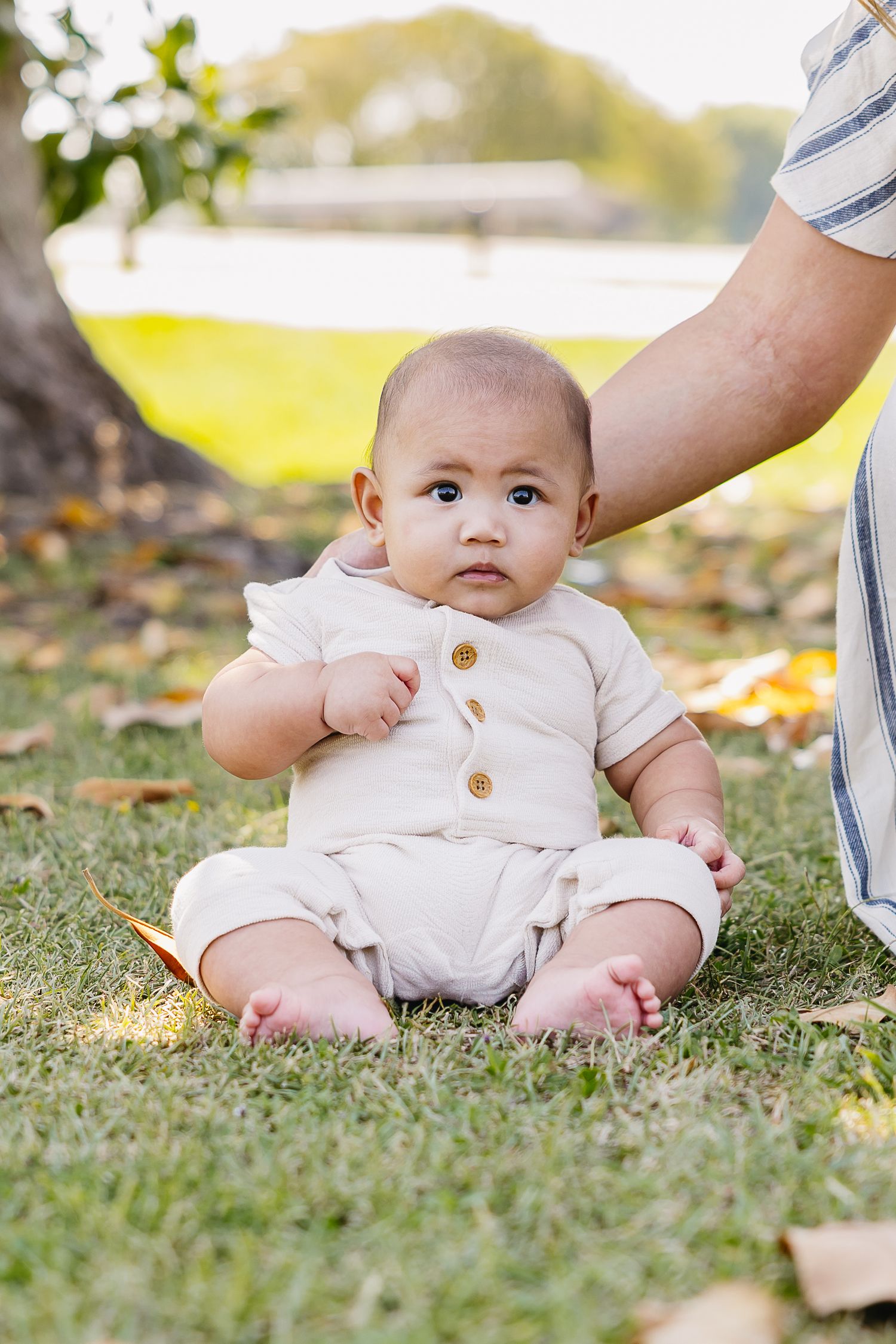 Adorable baby in cream outfit sitting on green grass in a sunny park.