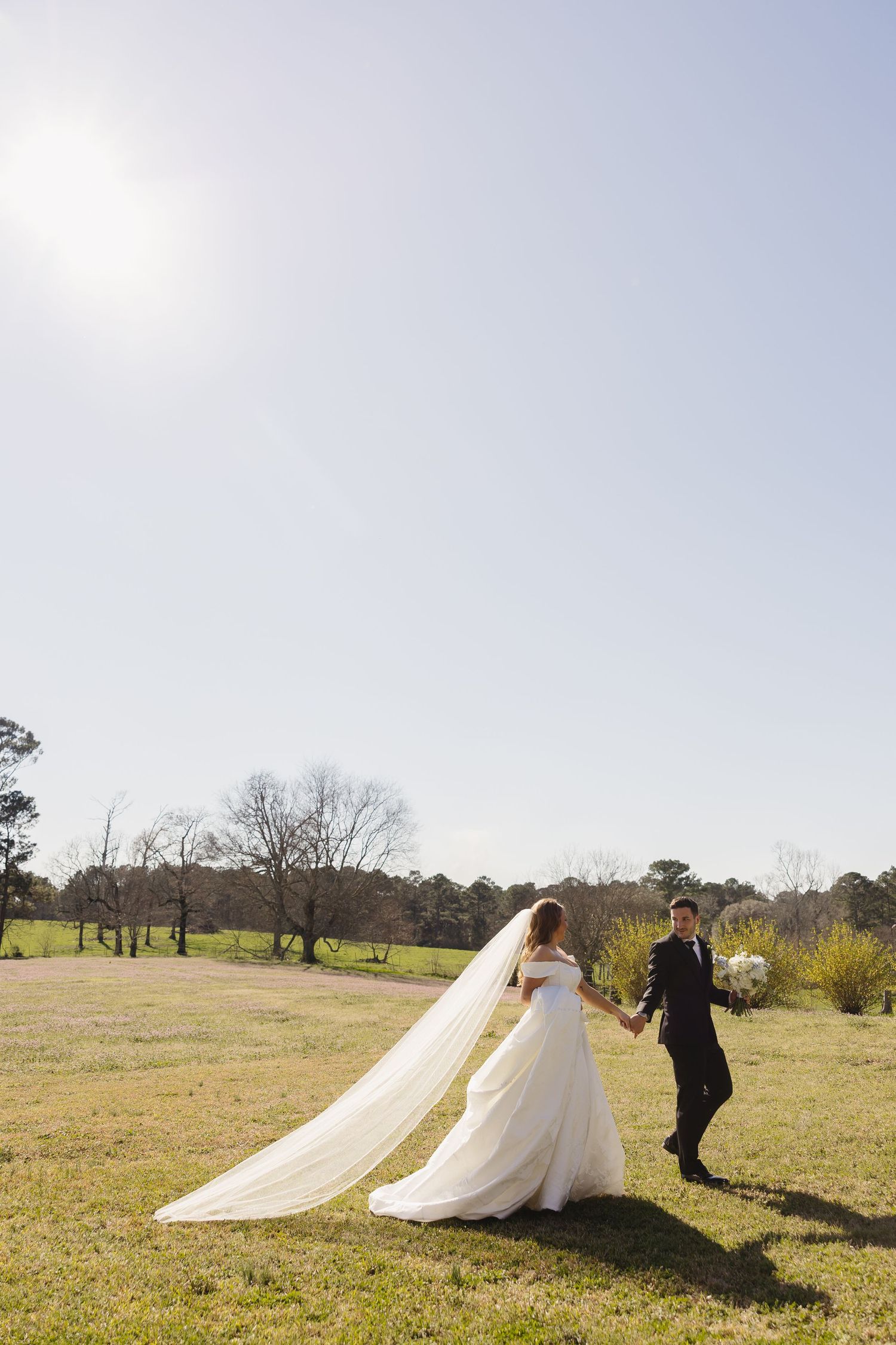 Bride and groom holding hands and walking across a sunny open field, veil trailing behind gracefully.